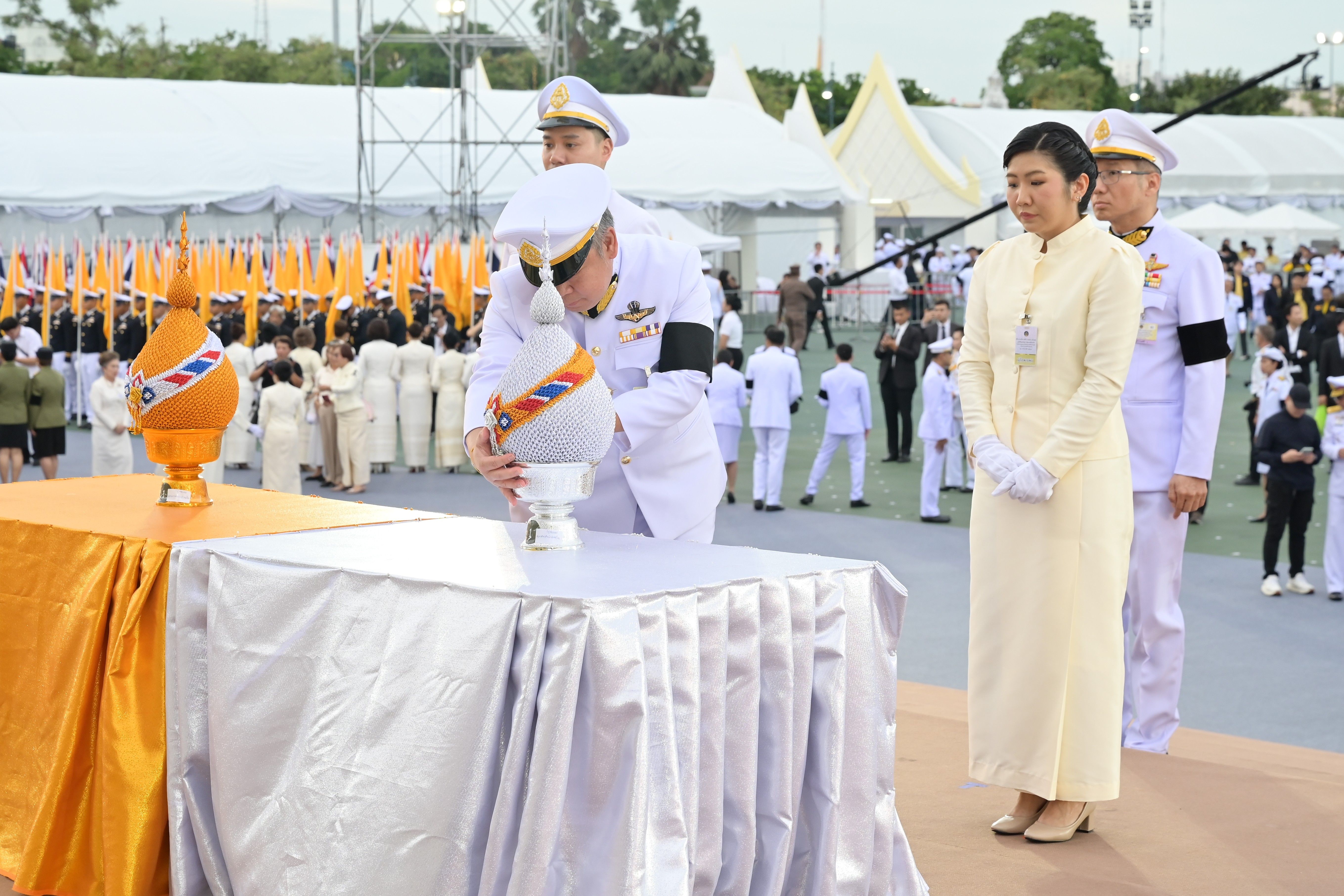 Man in white uniform pours from ornate orange urn during a solemn outdoor ceremony.