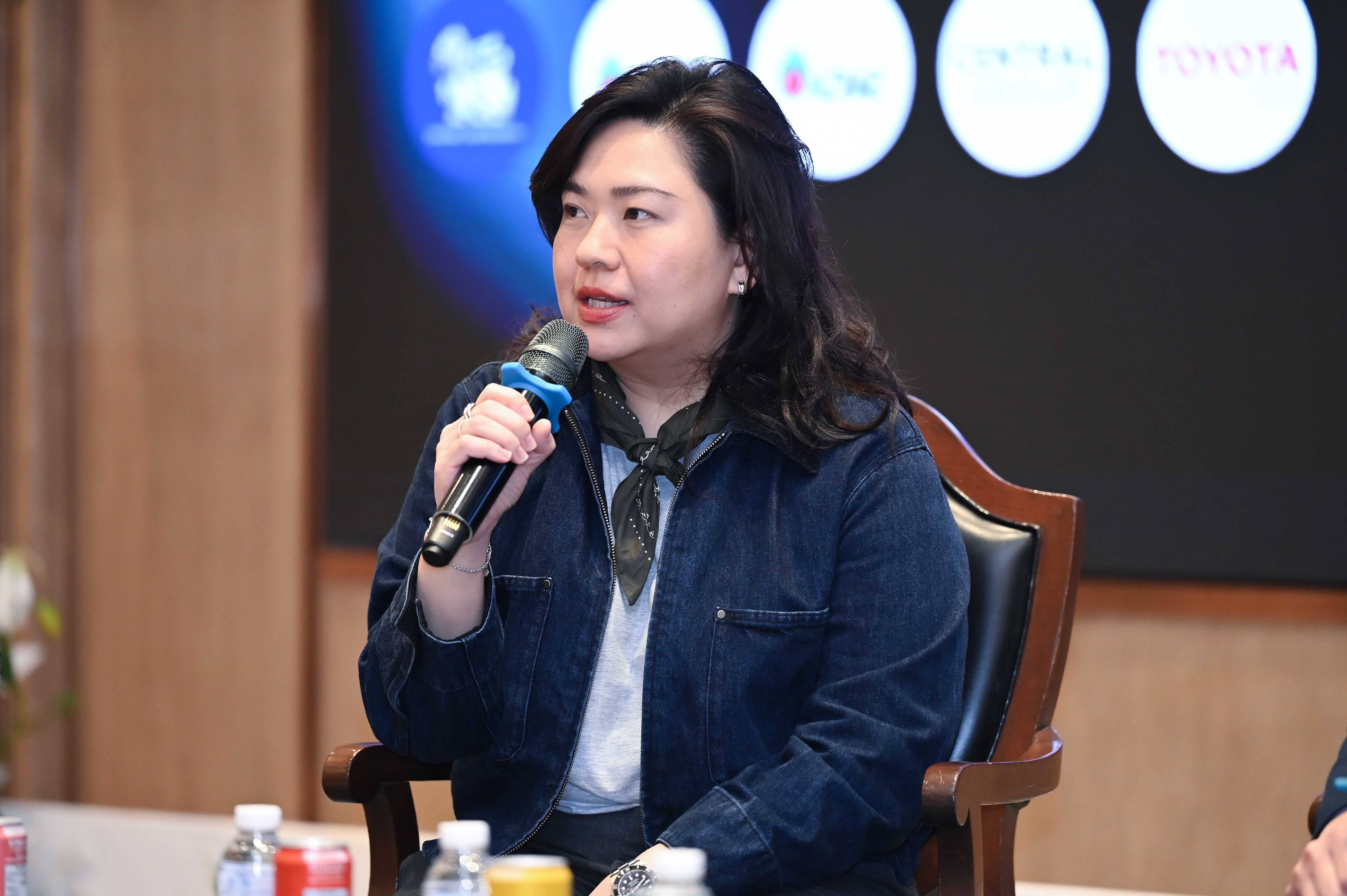 A woman in a denim jacket speaks into a microphone at a conference.