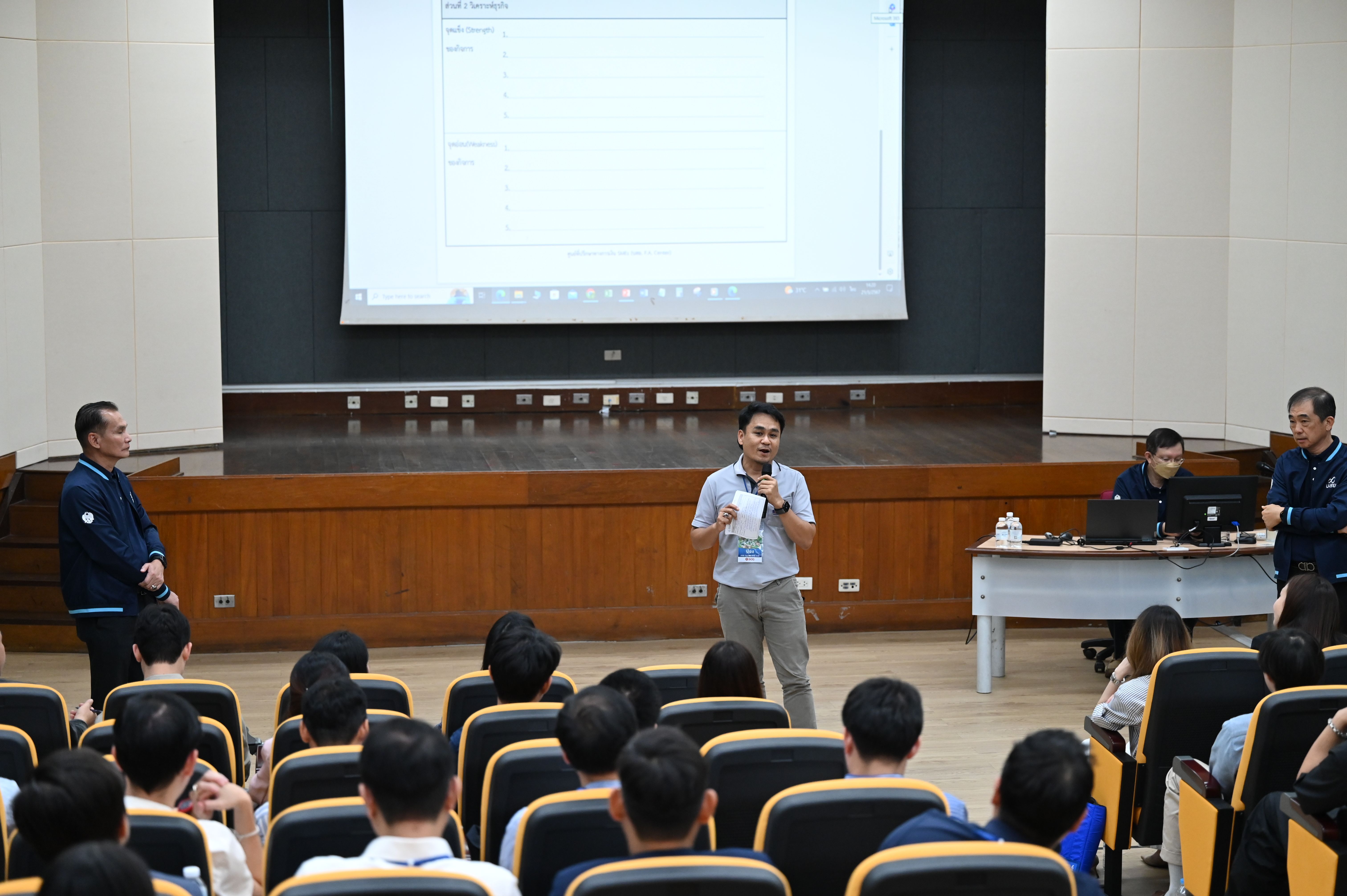 A male speaker addresses an audience from a stage, with a large projector screen displaying content.