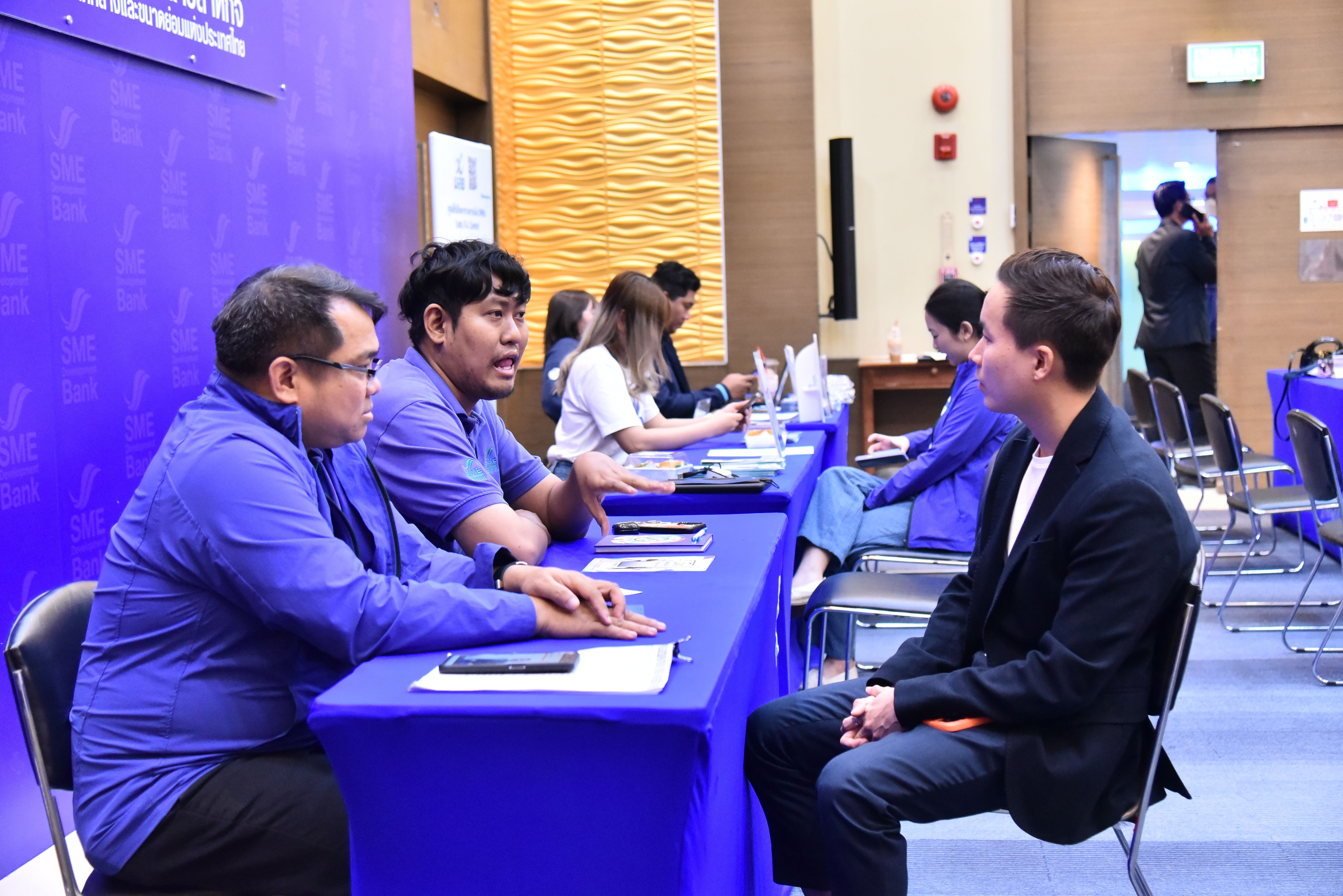 Two men in blue shirts interview a candidate at a blue table in a professional setting.
