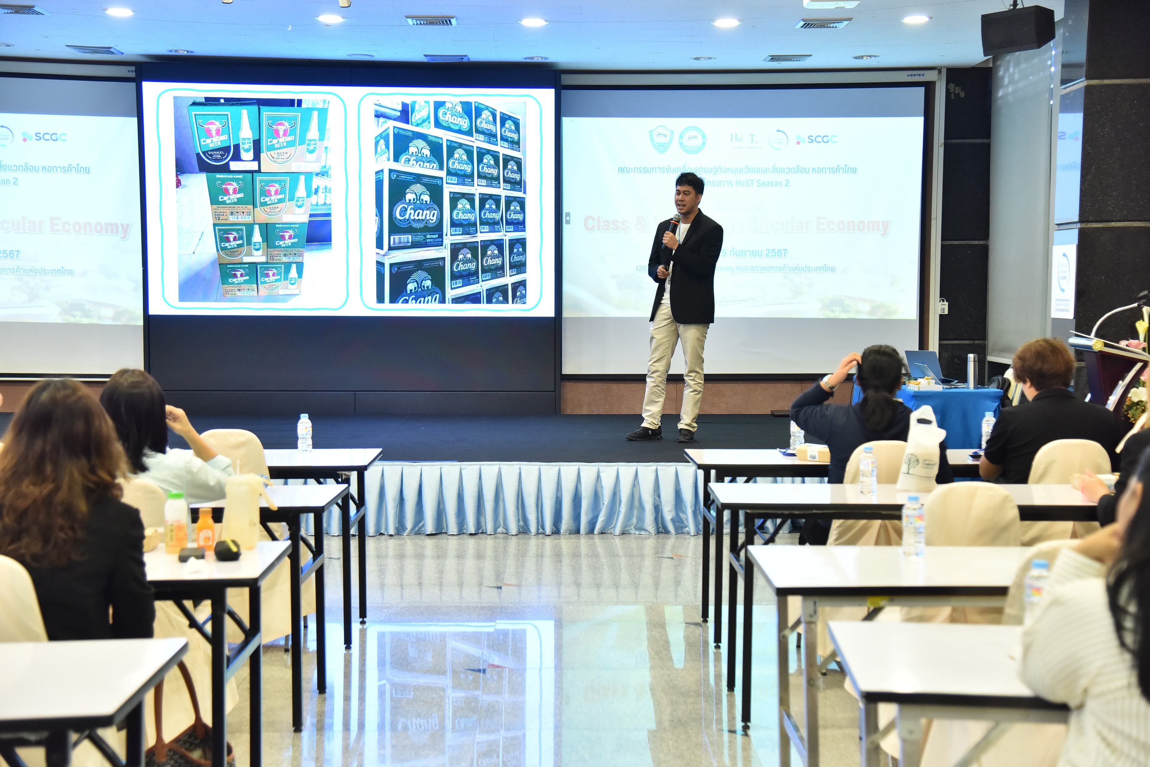 Man presents product packaging on large screens to an audience in a classroom setting.