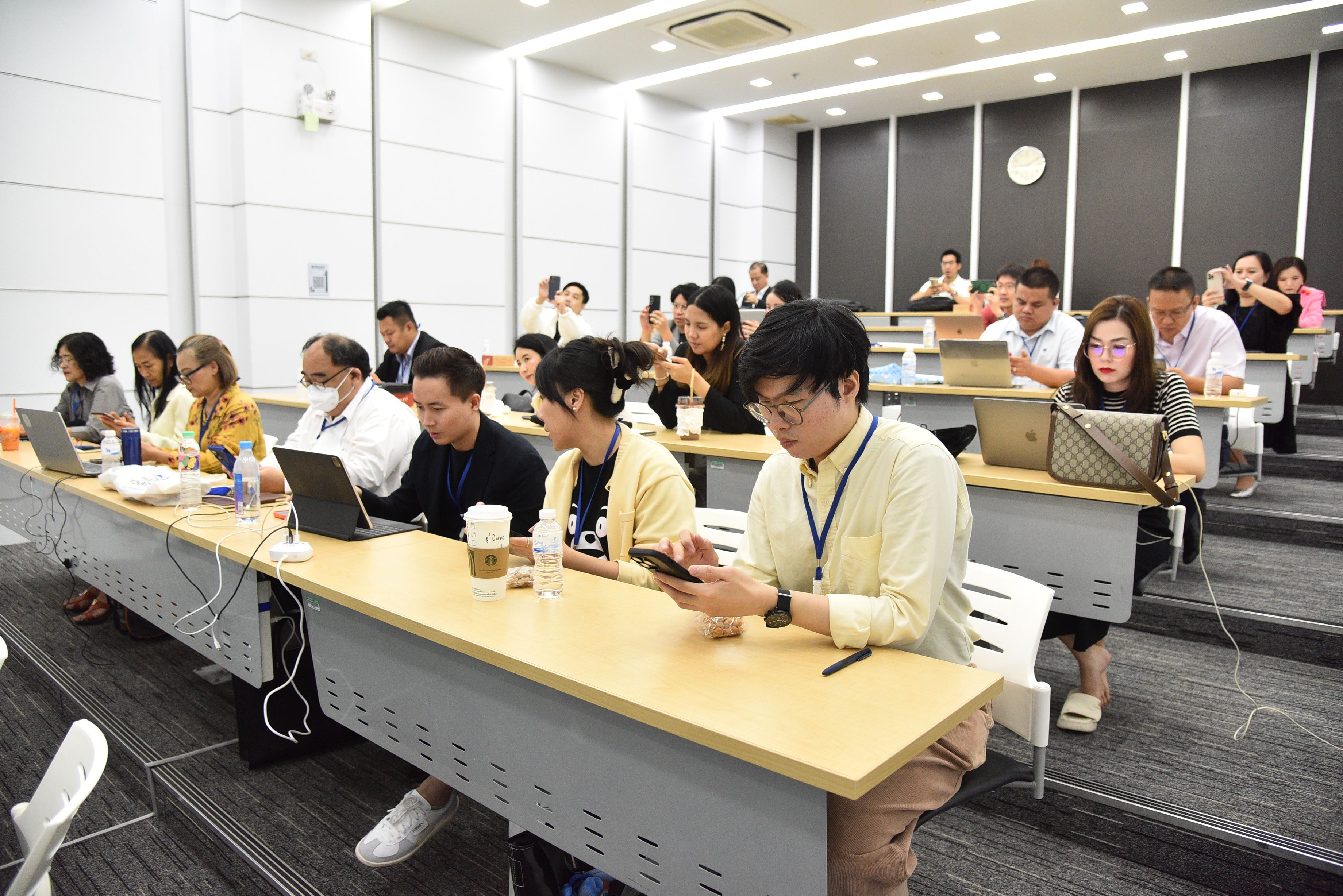Adults sit at desks in a brightly lit classroom, many using laptops and phones.