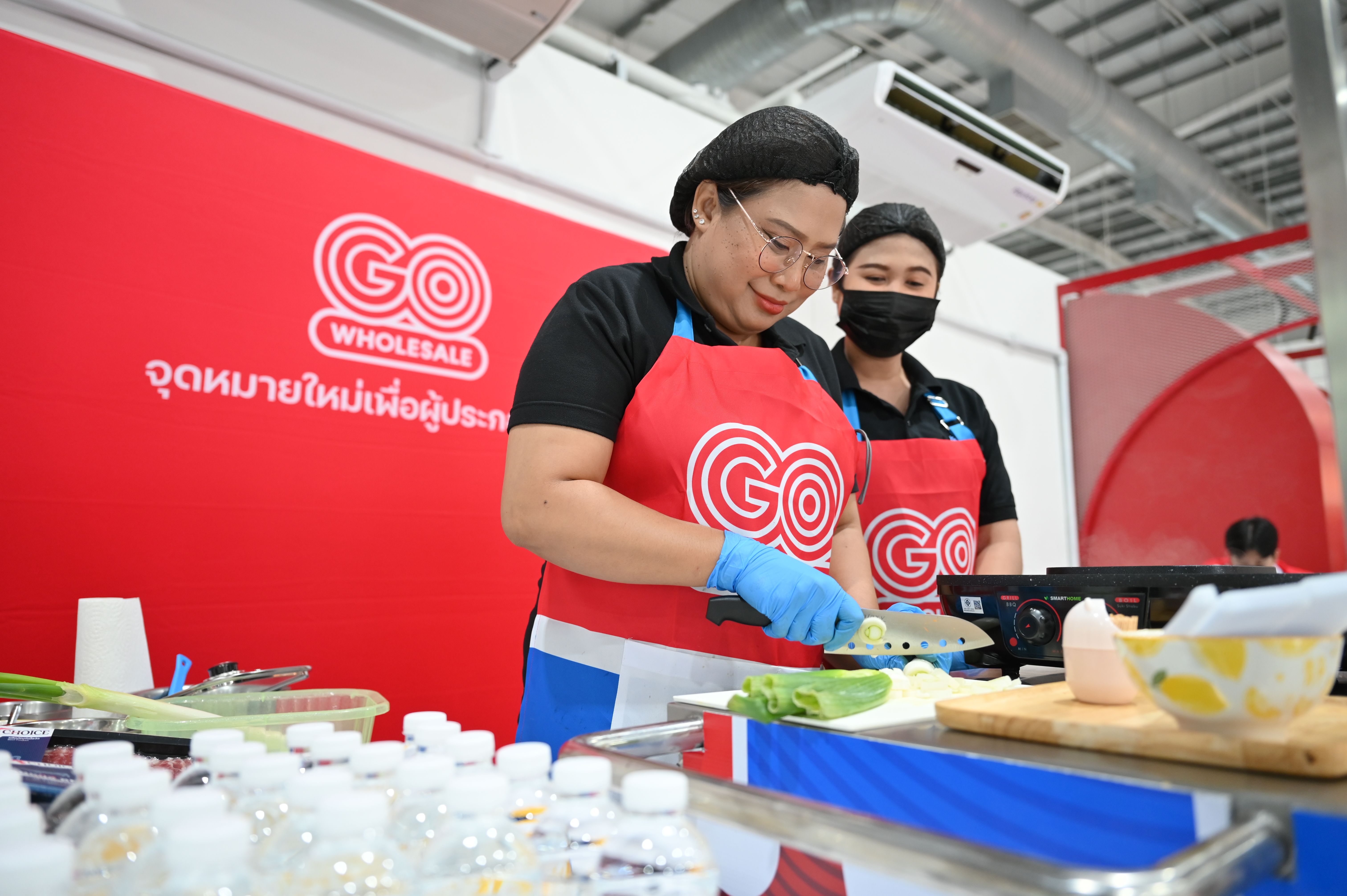 Two people in GO WHOLESALE aprons preparing food at a cooking station.