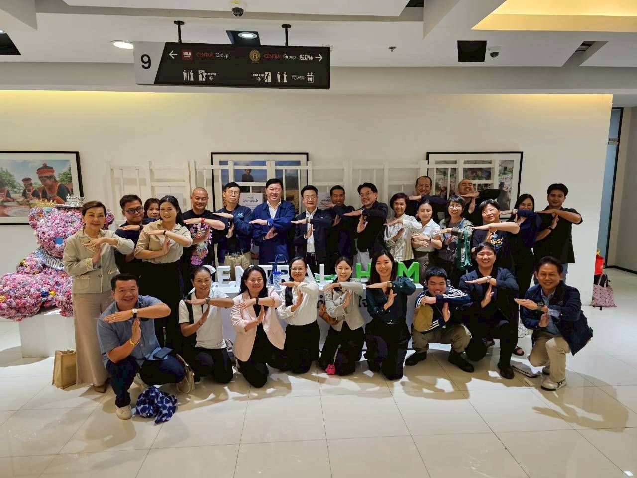 A large group of smiling people performing a unified hand gesture in a modern indoor setting.