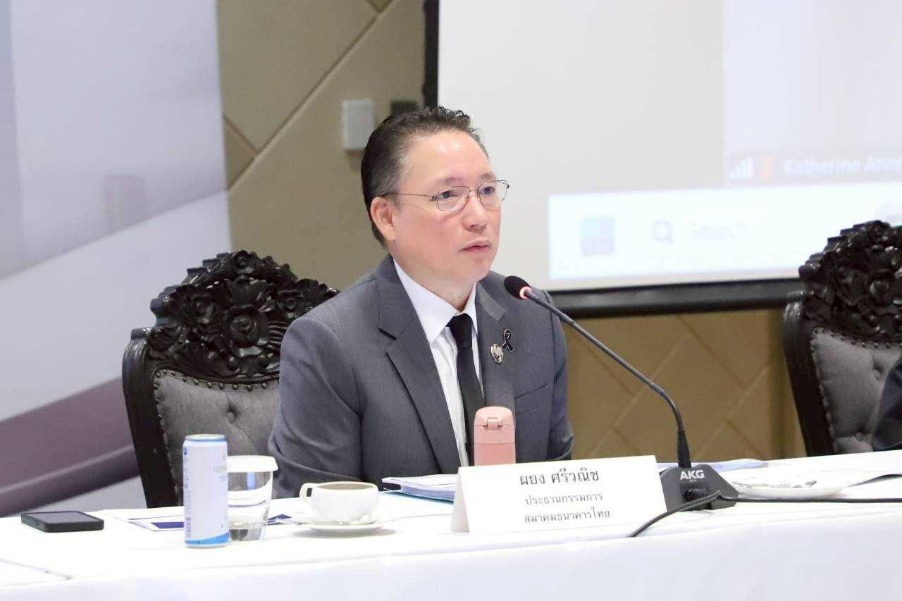 A man in a suit and glasses speaks into a microphone at a formal meeting table.