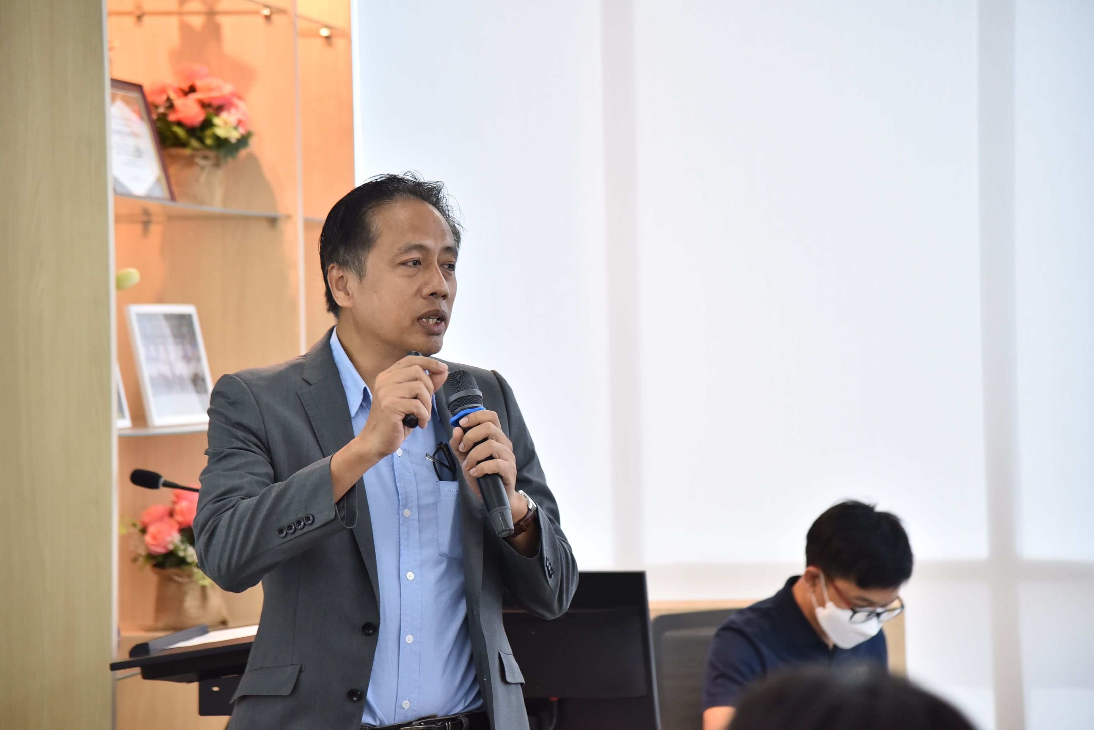 Man in a grey suit speaking into a microphone at an event.