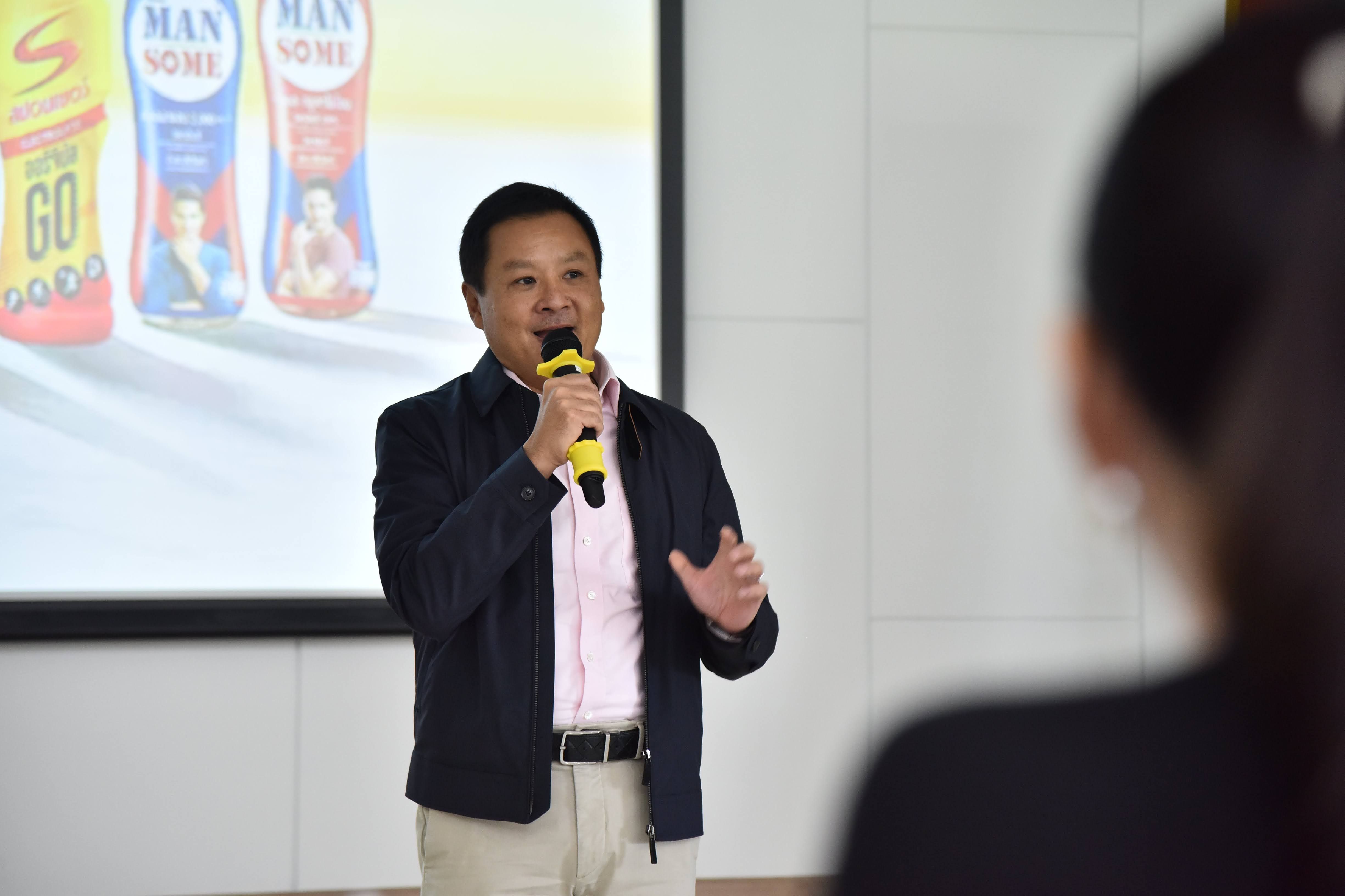 Man in a dark jacket speaking into a microphone during a presentation with product bottles shown.