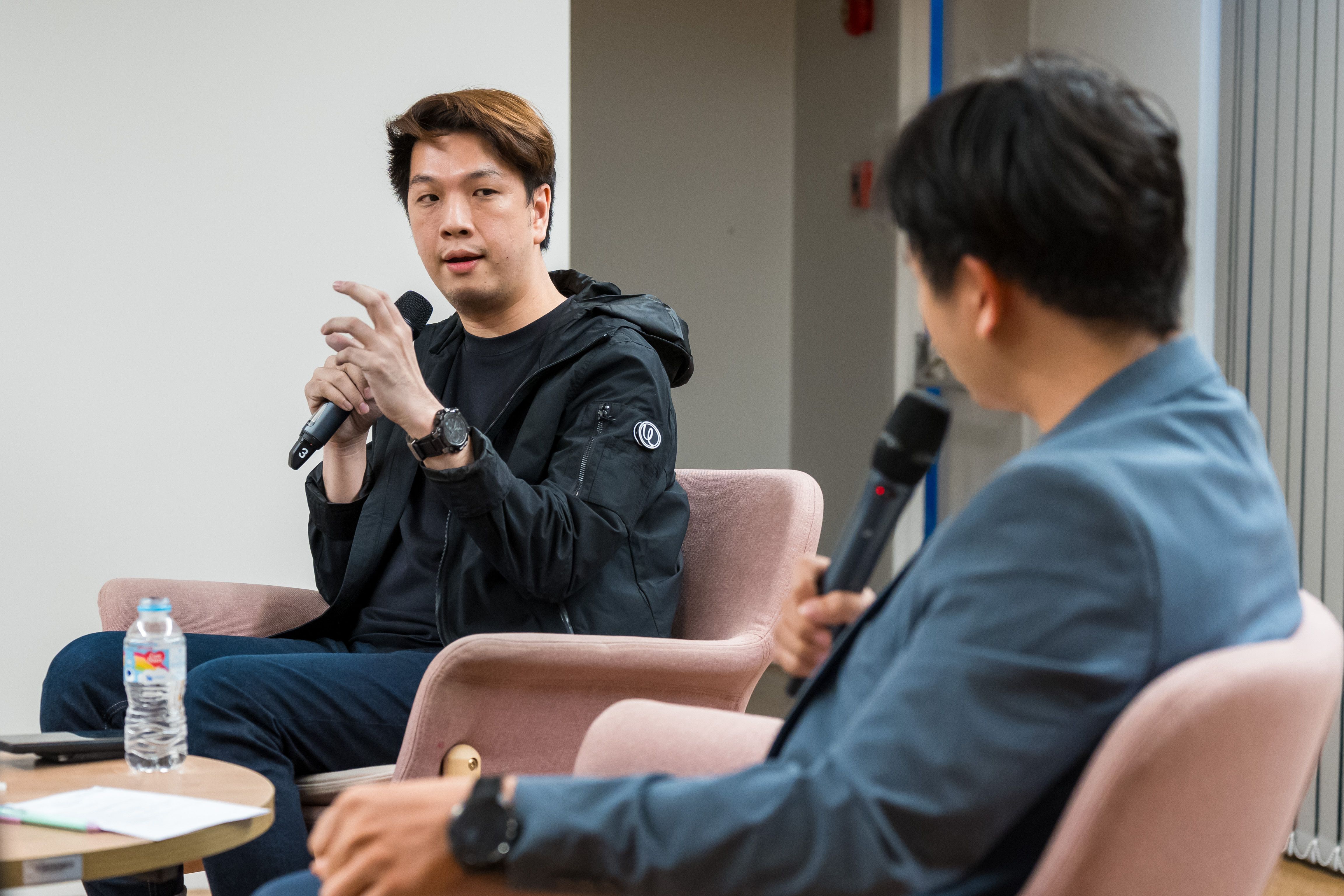 Man in dark jacket speaks into microphone, gesturing during an interview with another man.