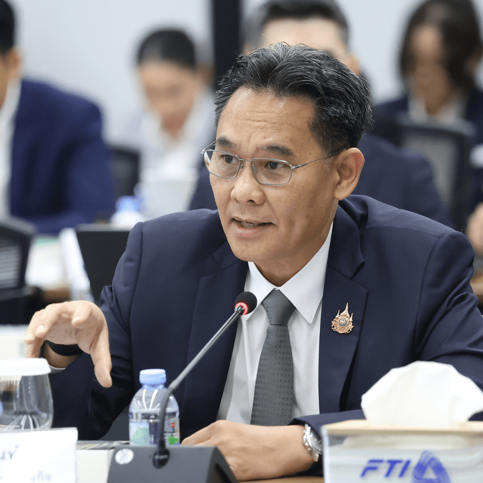 An Asian man in glasses and a dark suit speaks and gestures at a meeting table.