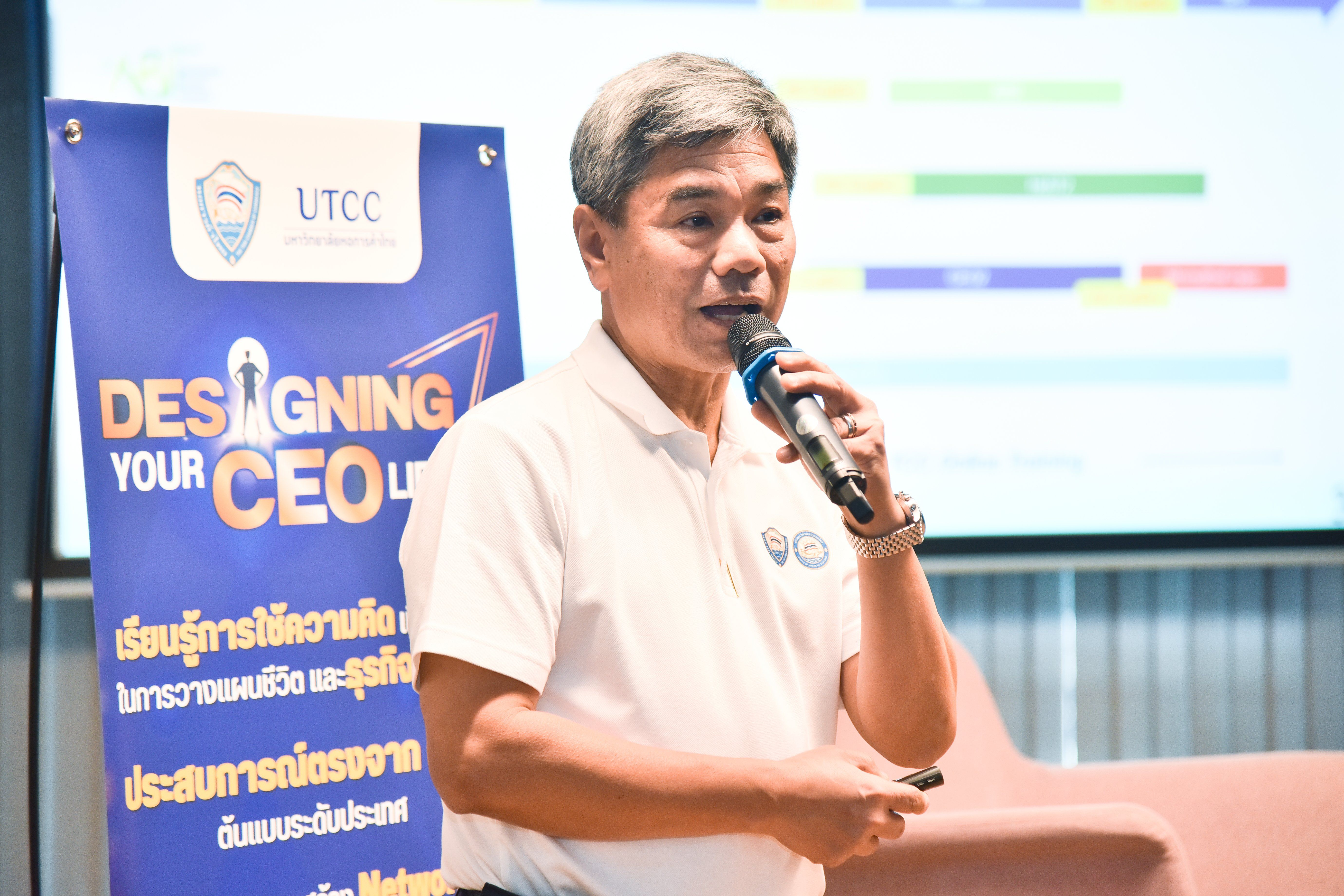 Man in white shirt speaks into microphone near "Designing Your CEO" banner and screen.