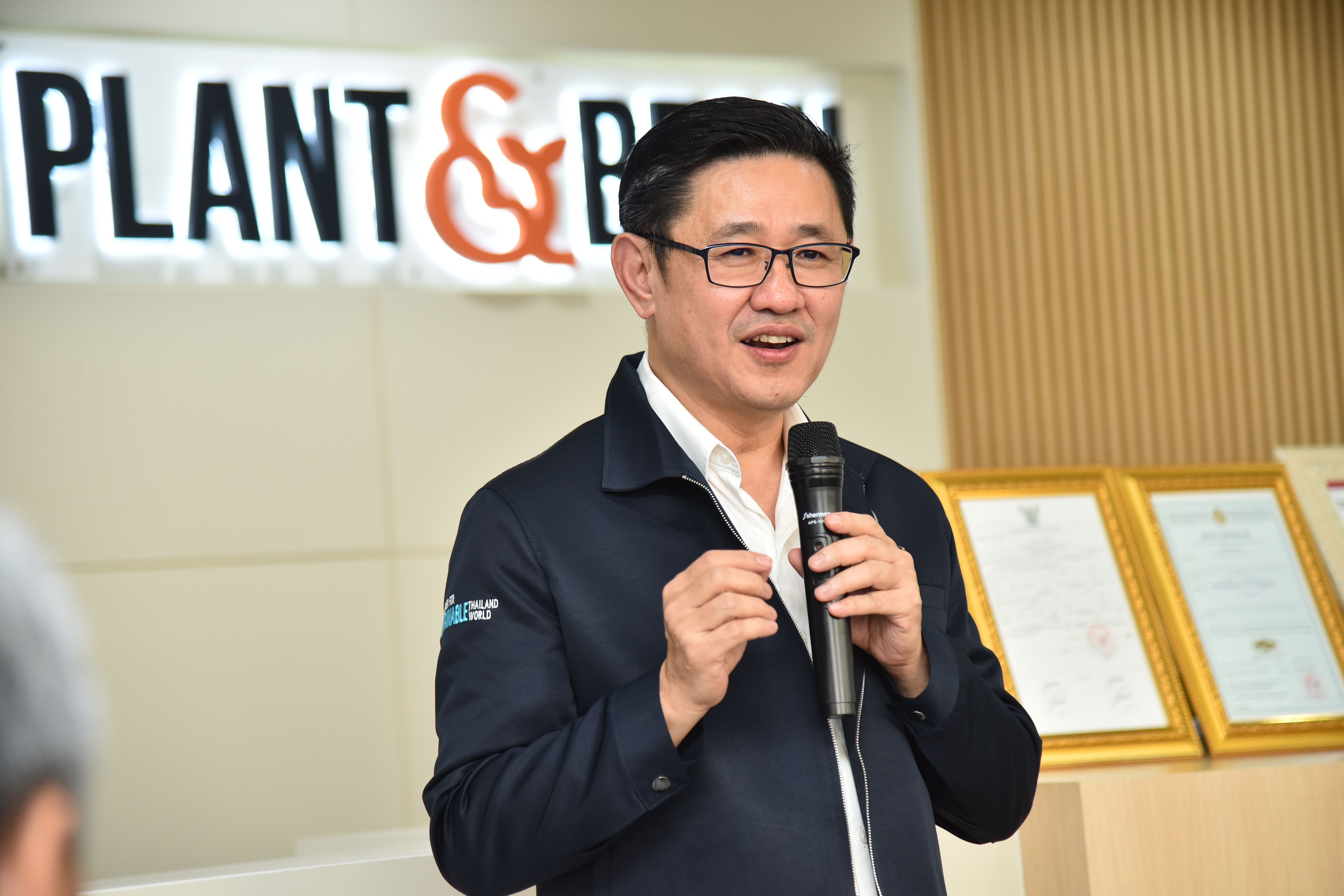 An Asian man speaks into a microphone, with a 'PLANT &' sign behind him.