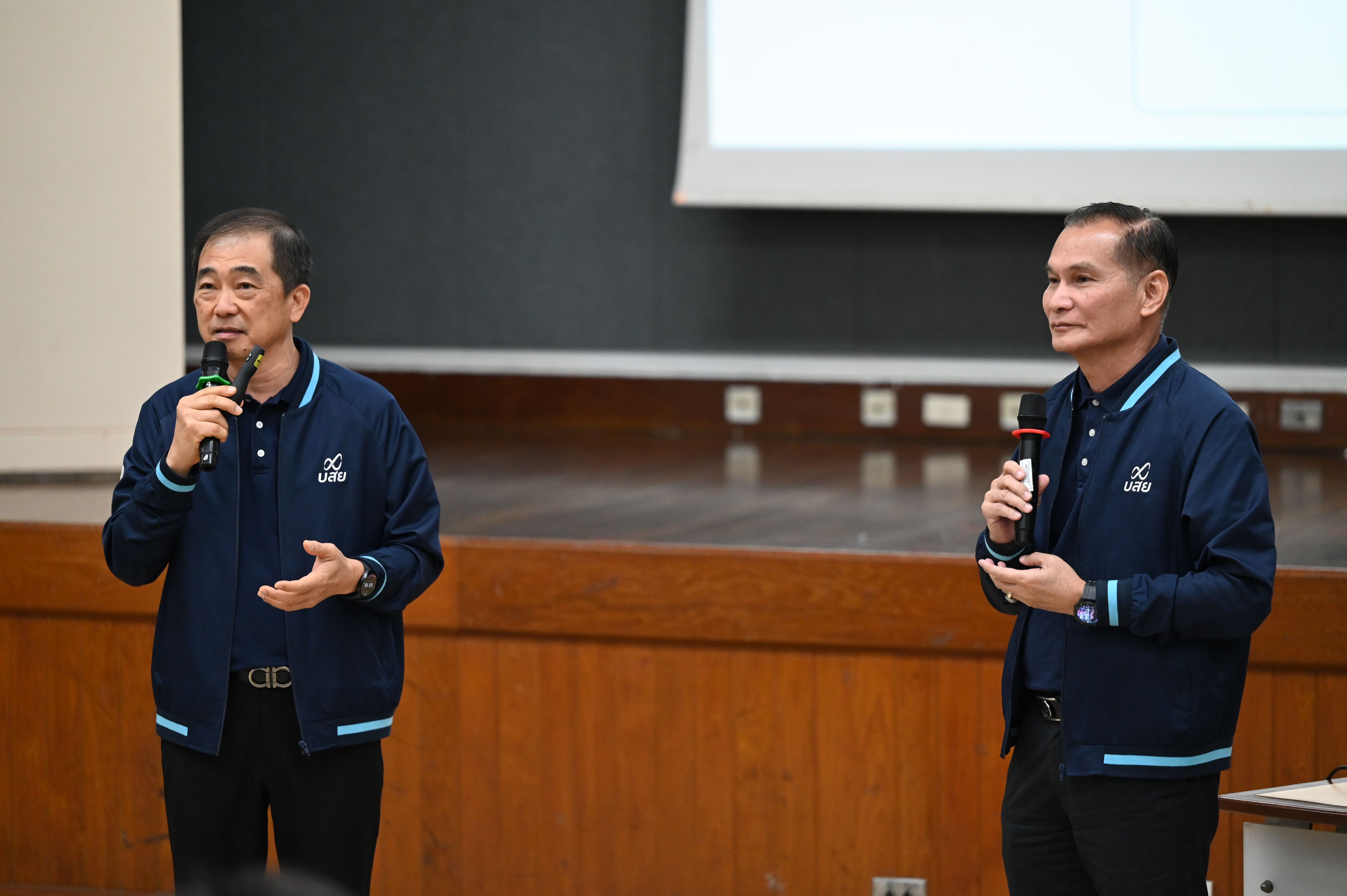 Two men in matching blue jackets speaking into microphones on a stage.