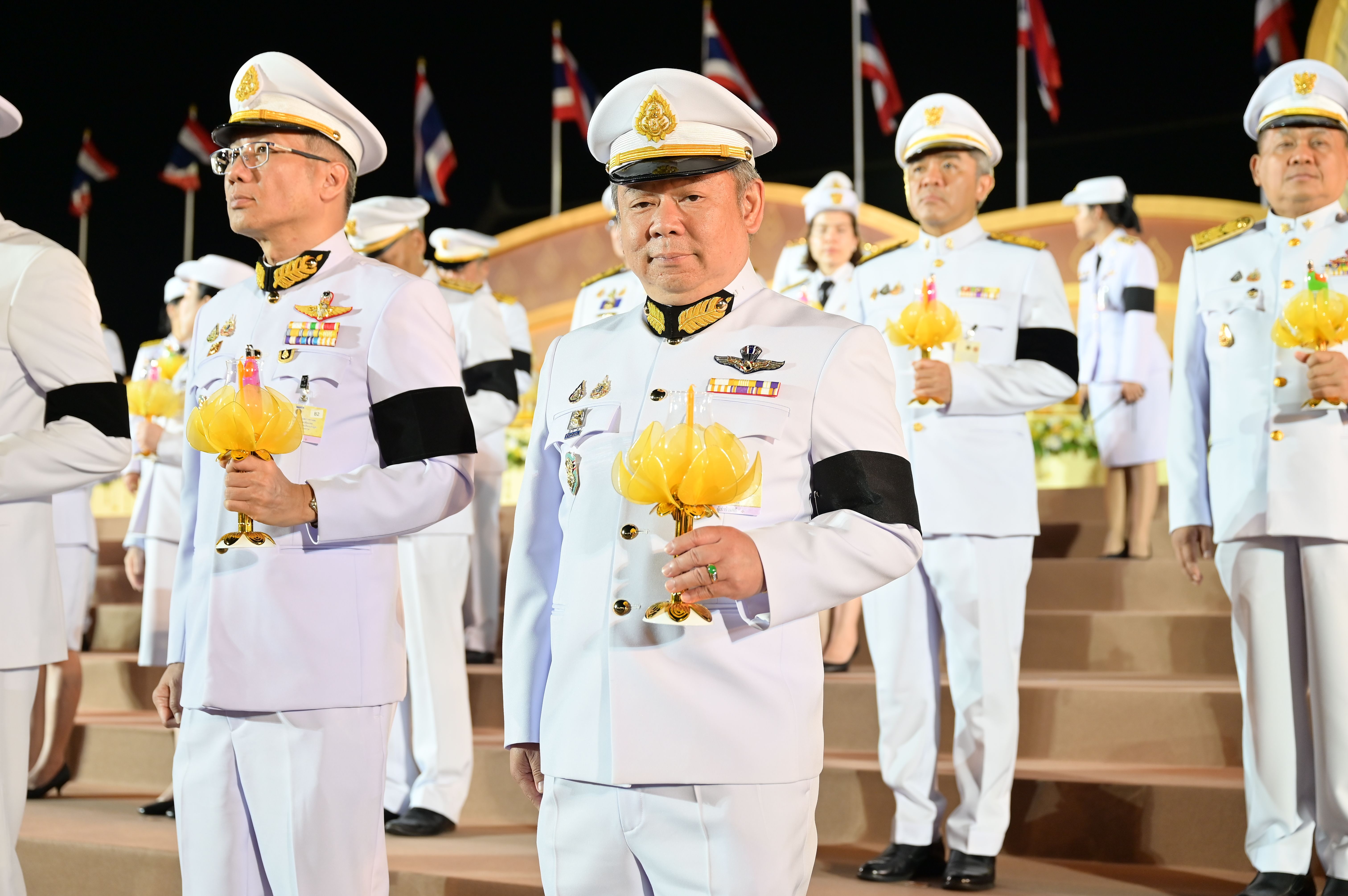 Officials in white uniforms with black armbands hold yellow ceremonial objects at a night ceremony.