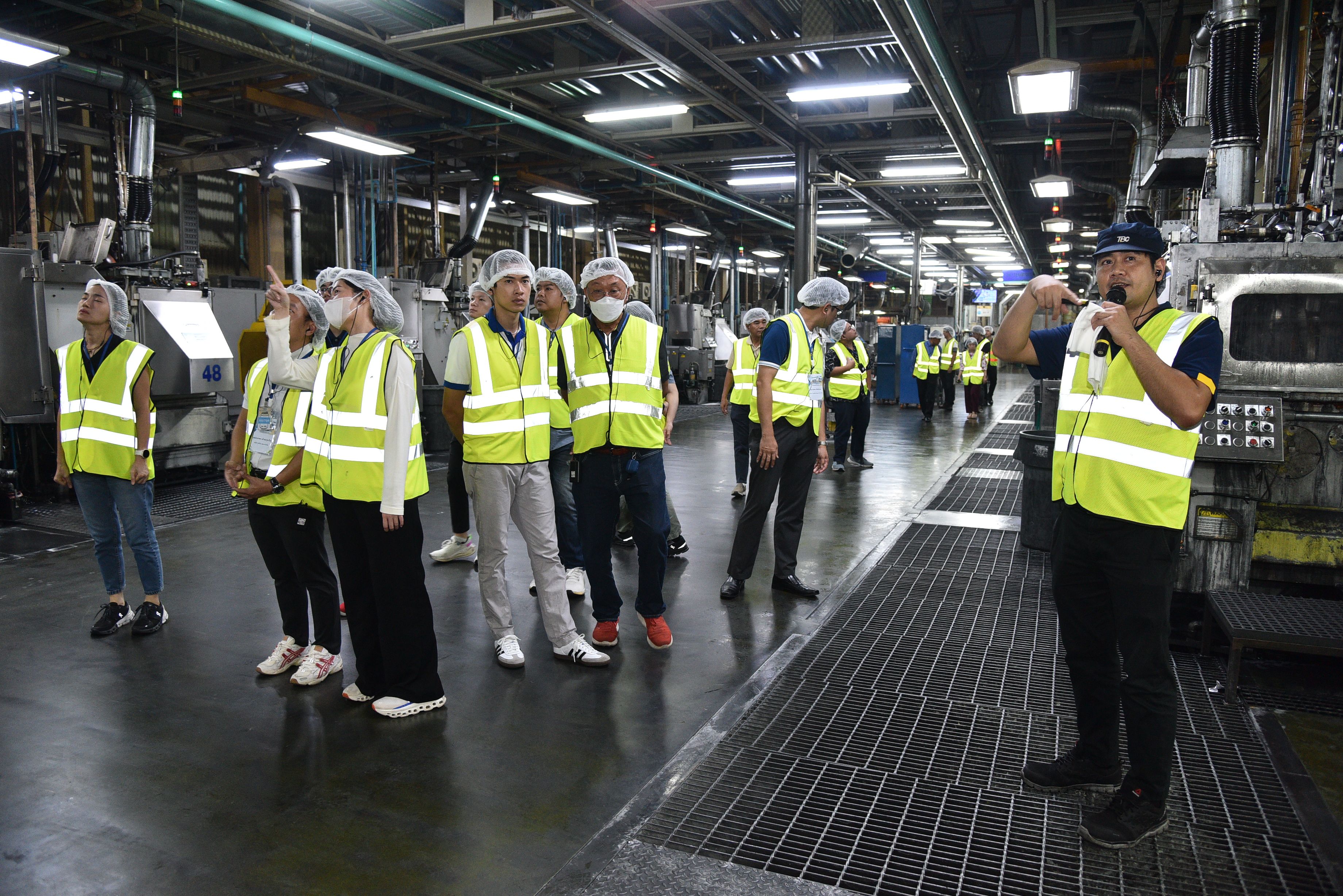 Guide addresses visitors in safety vests and hairnets during a factory tour.