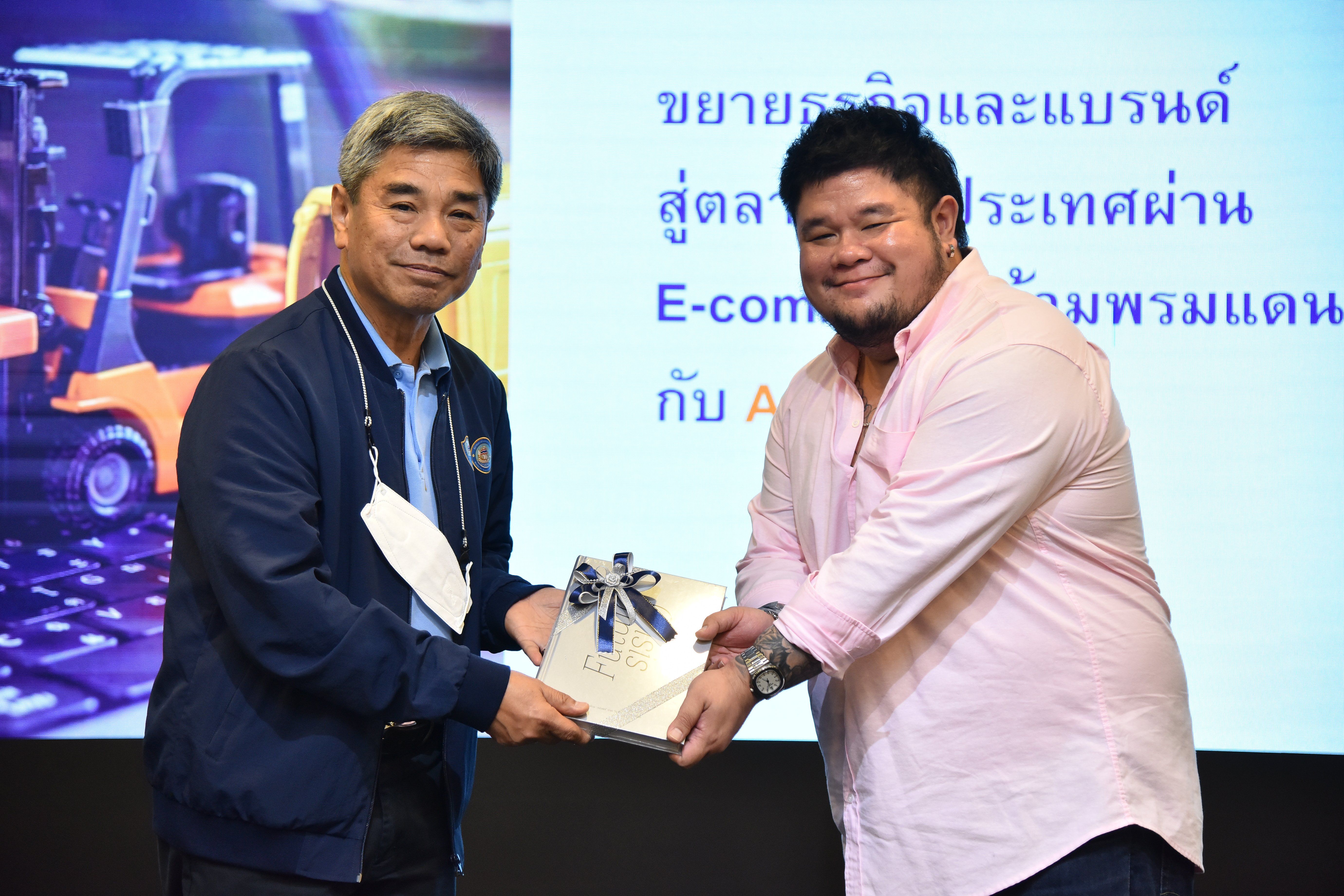 Two men exchange a wrapped gift on stage with a screen displaying Thai text.