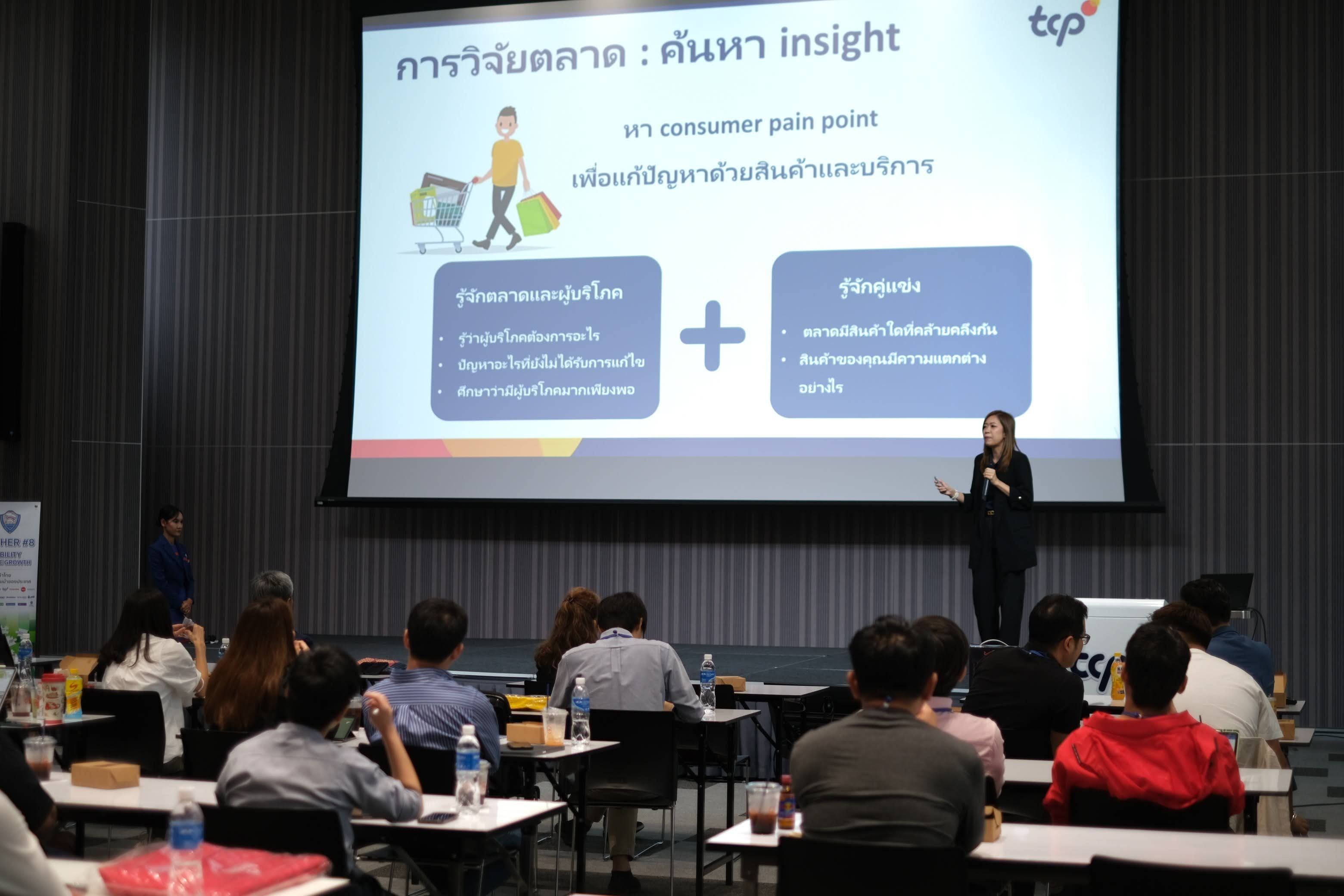 A woman presents to an audience with a Thai presentation screen displaying a diagram.
