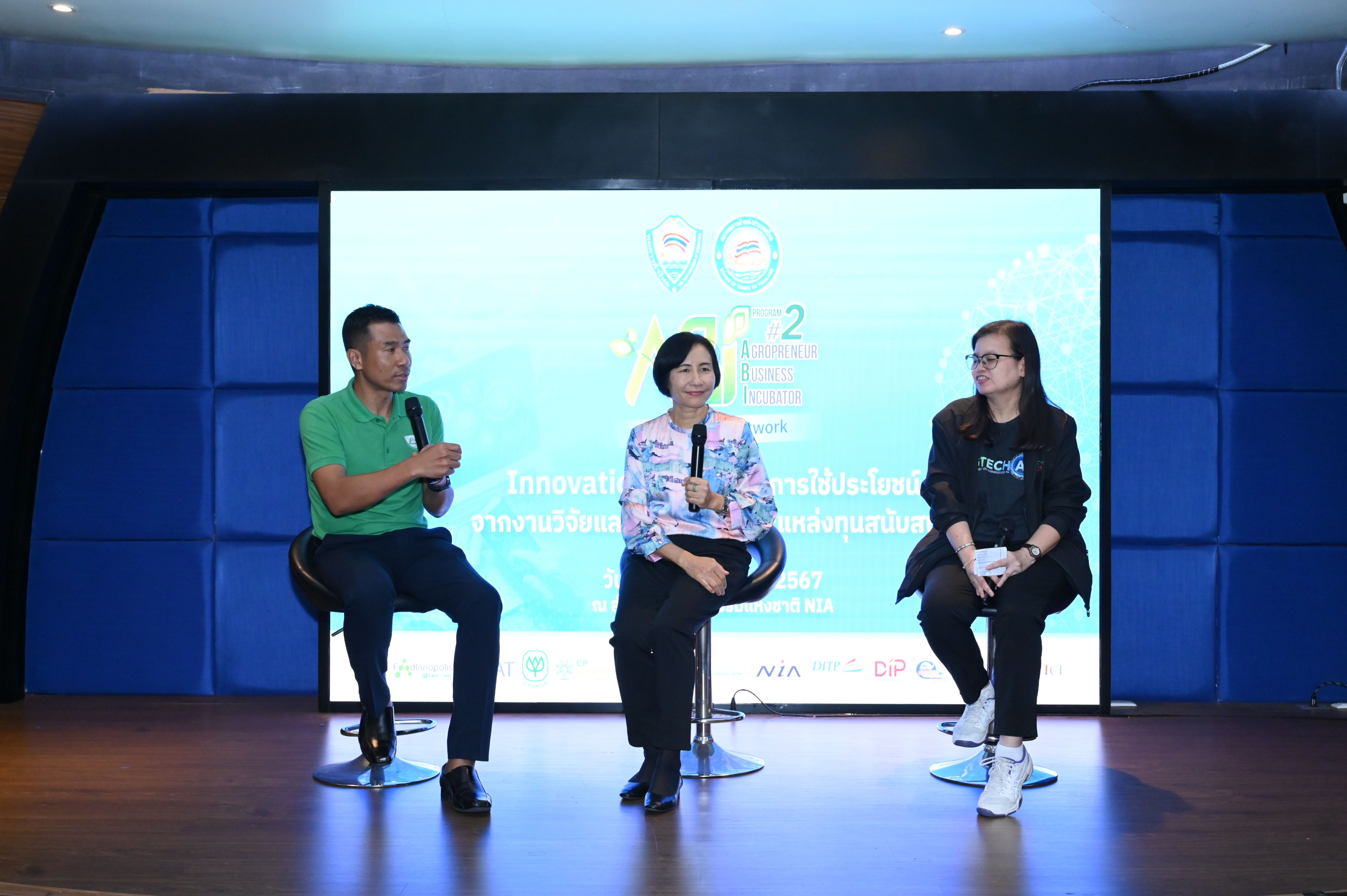 Three panelists discuss on stage during a conference with a large screen behind them.