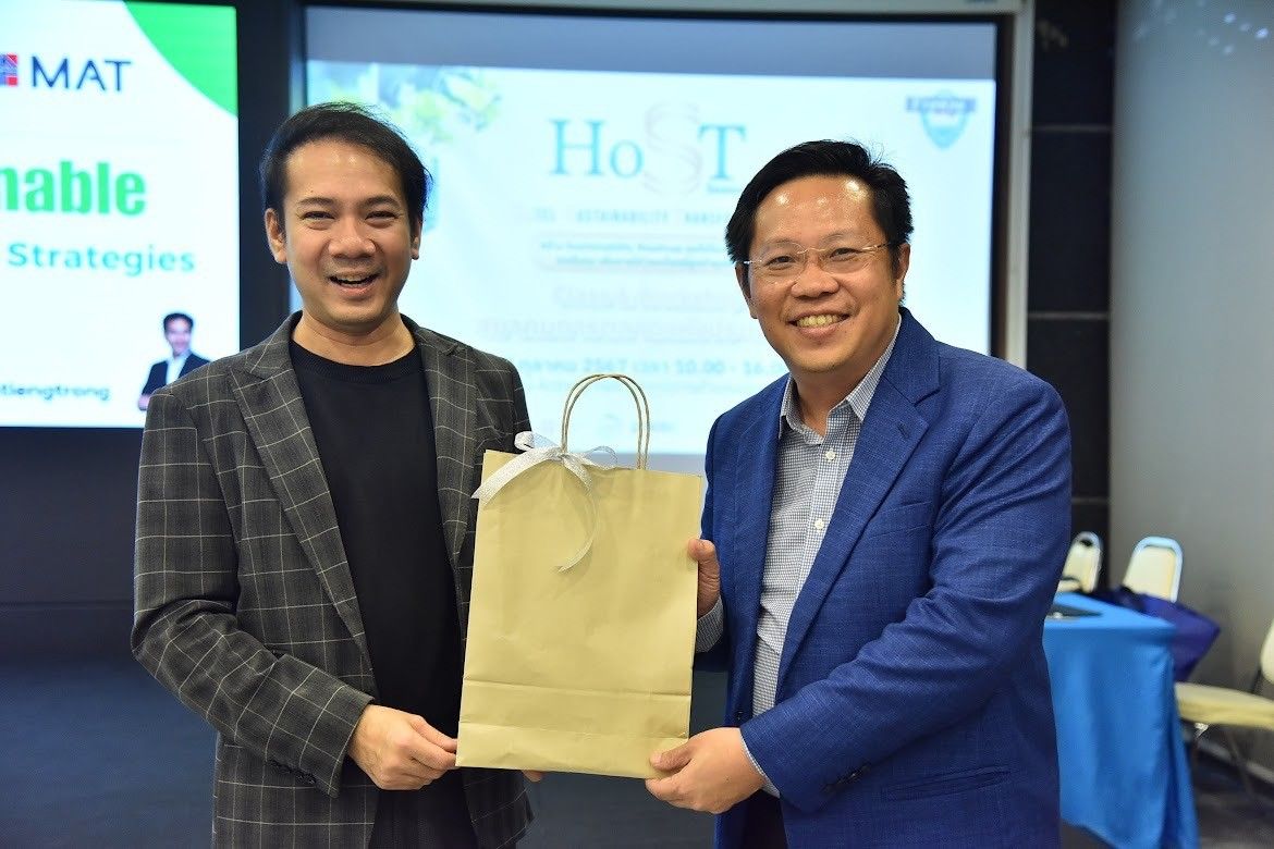 Two smiling men at a professional event jointly hold a brown paper gift bag.