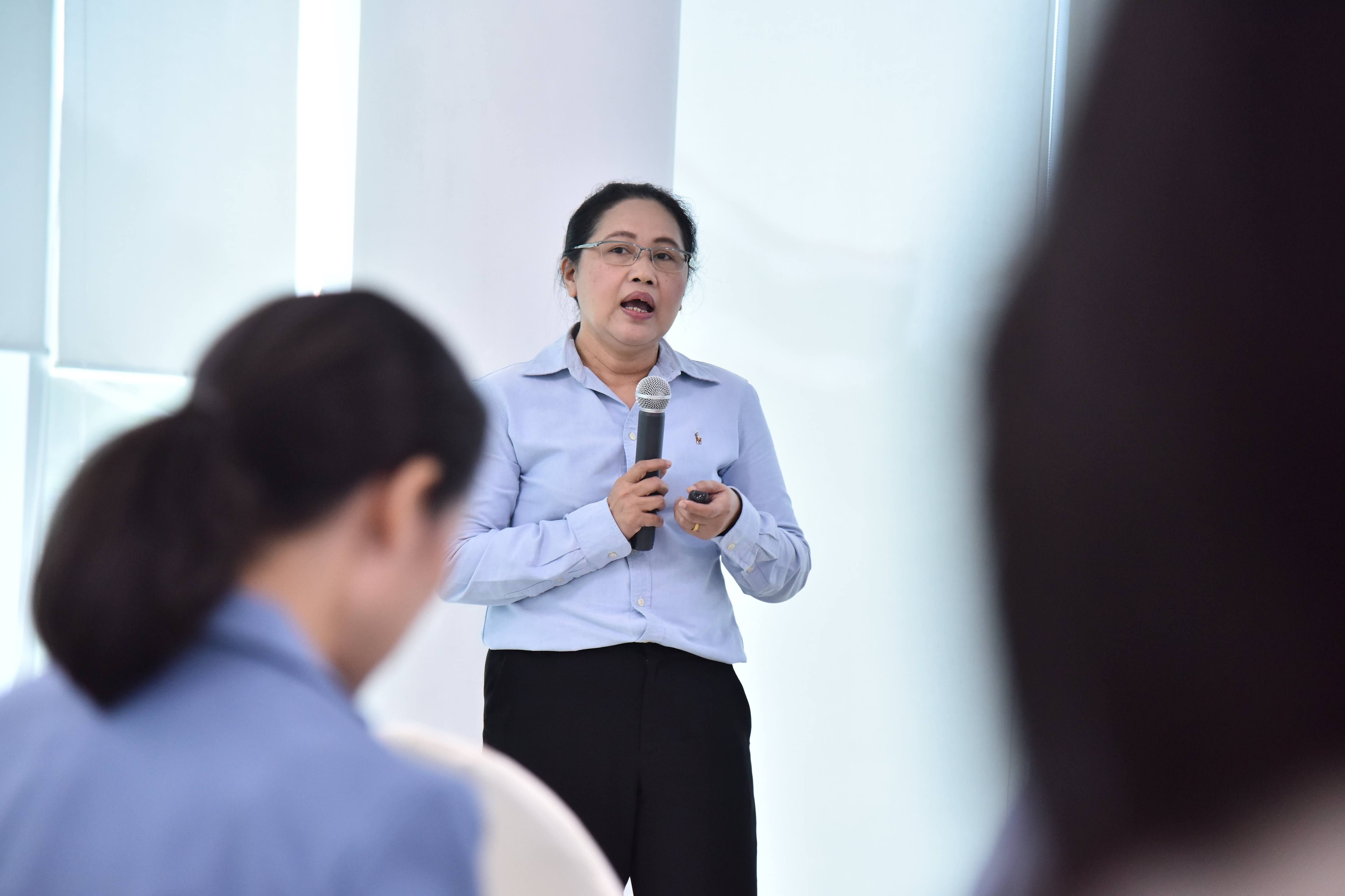 Woman in blue shirt speaking into microphone, addressing an audience indoors.