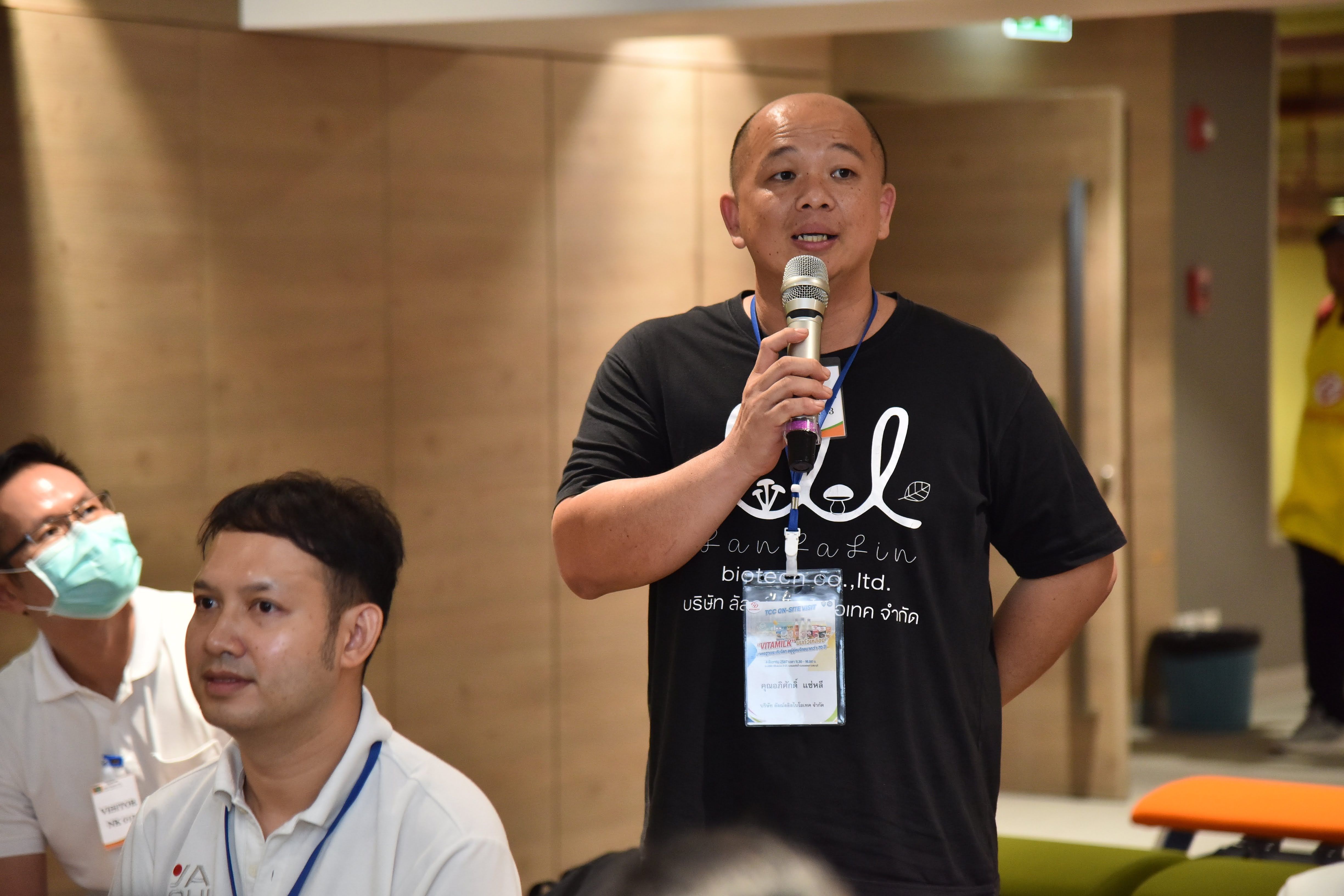Man in black t-shirt speaks into microphone at an indoor event.