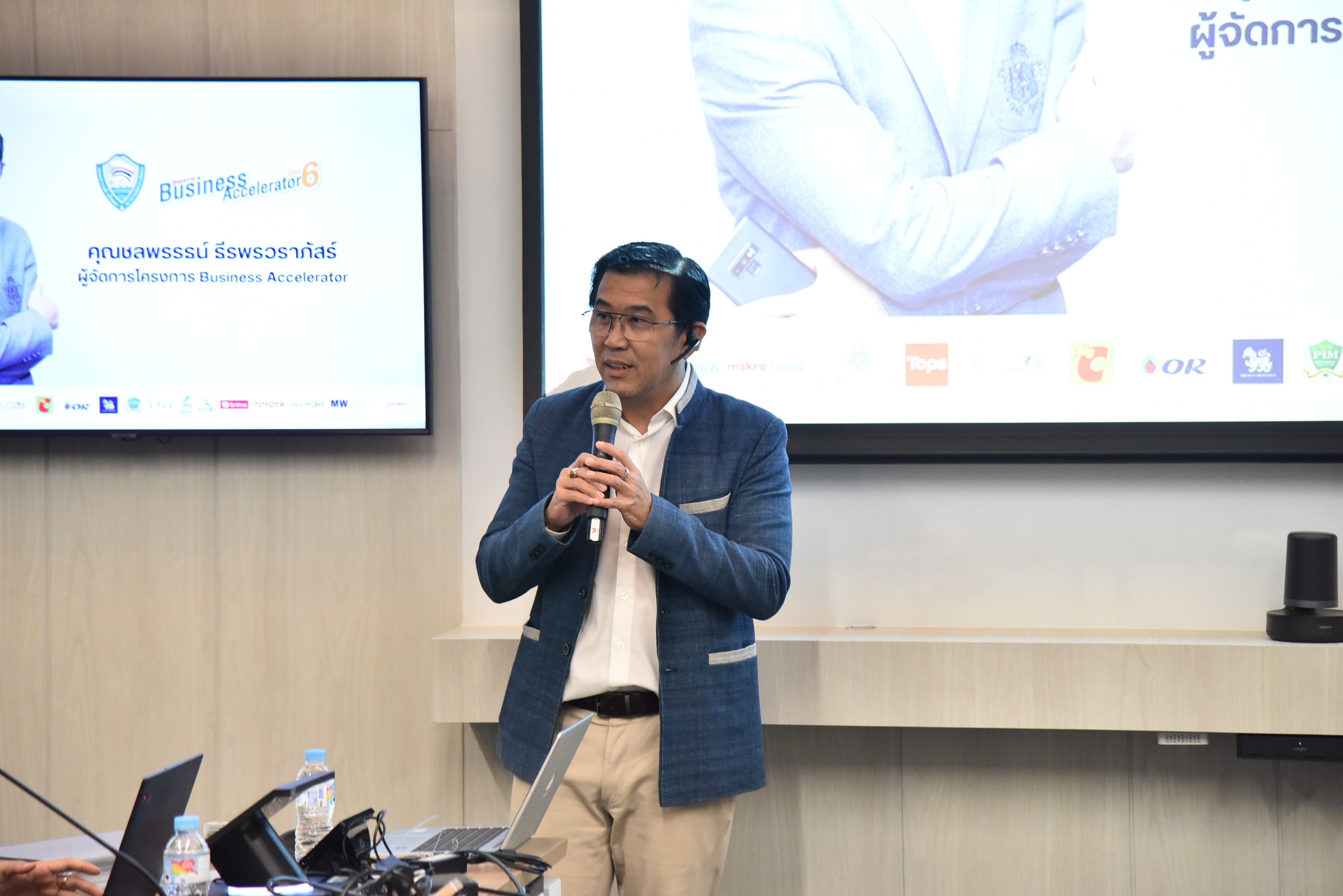Man in a blue blazer speaking into a microphone during a presentation in a conference room.