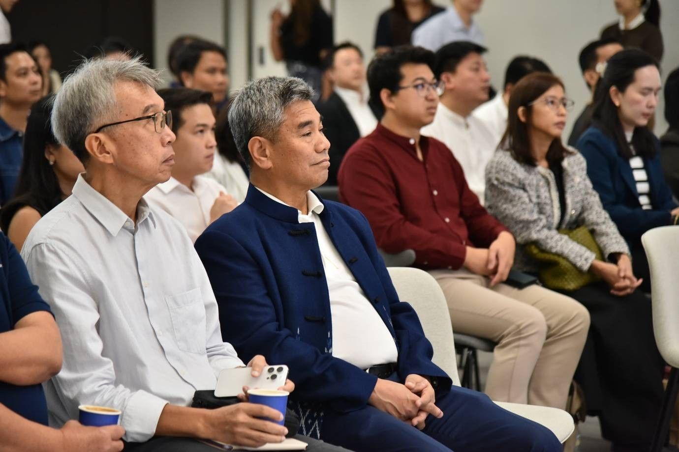 Two Asian men in the foreground attentively listen among a diverse audience at a professional event.