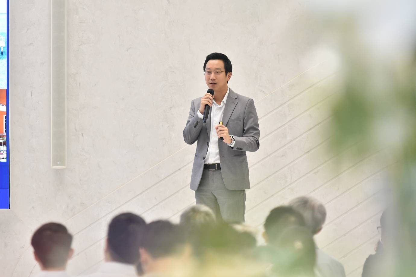 A man in a grey suit and glasses speaks into a microphone at an event.