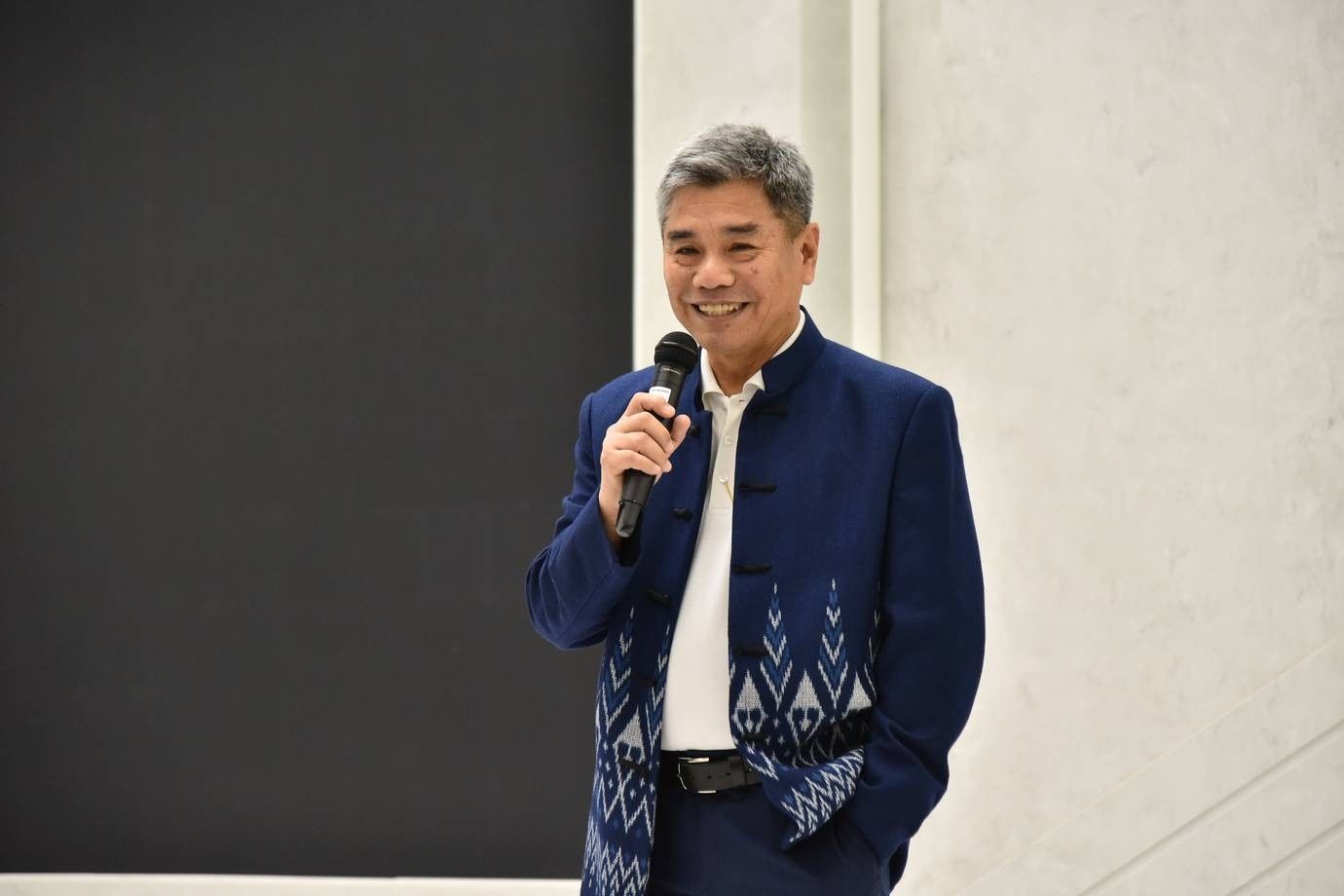 A smiling man in a blue patterned jacket speaks into a microphone.