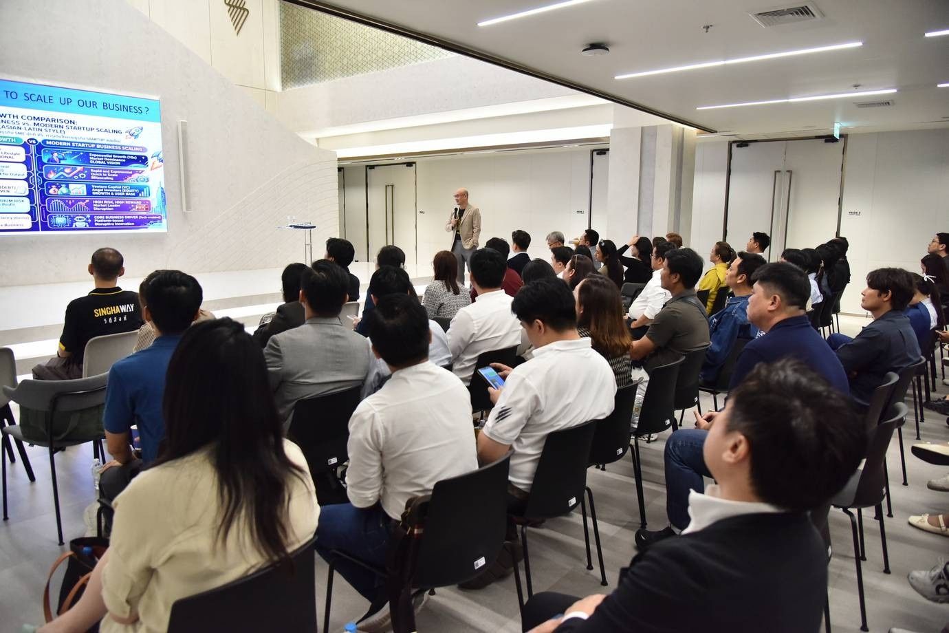 An audience watches a presenter with a data visualization on a large screen in a bright room.