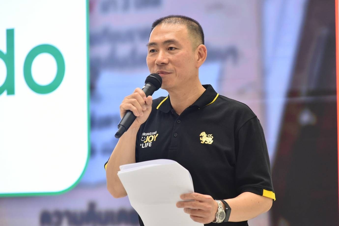 Man in black polo shirt with yellow logos speaking into a microphone.