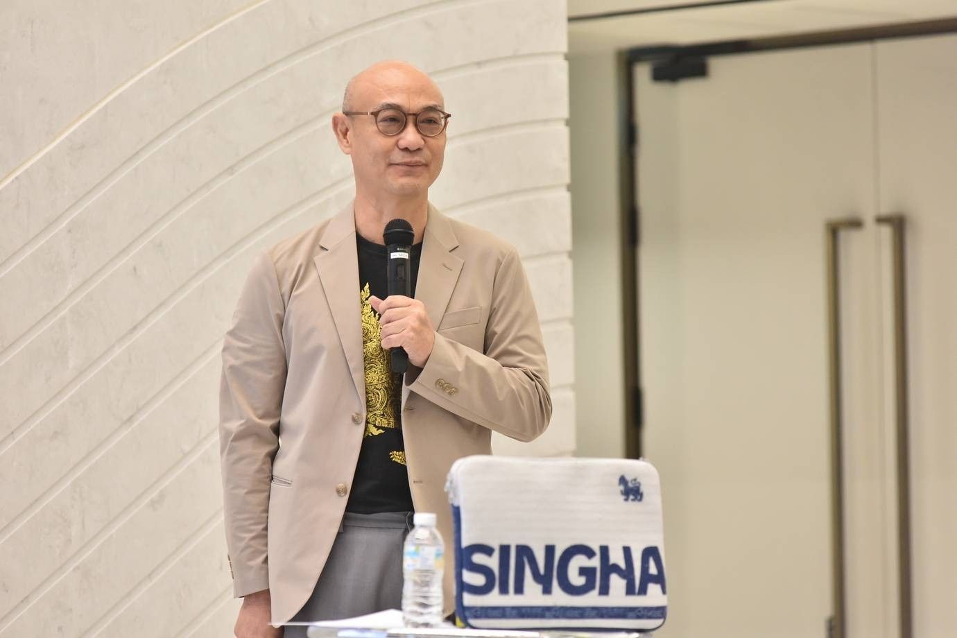 Bald man in glasses speaking into microphone next to a 'SINGHA' sign.