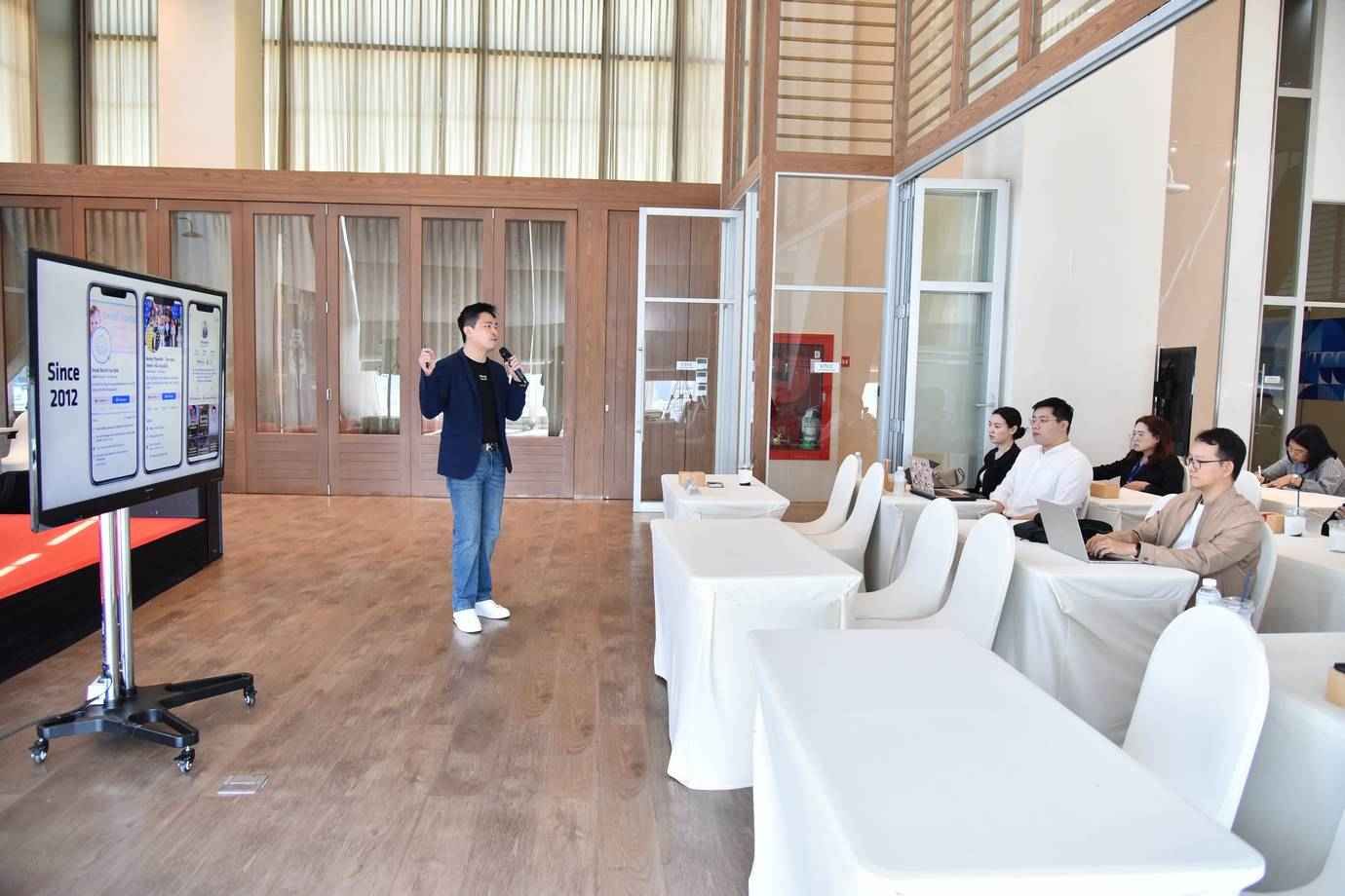 Man presenting to an audience with a microphone next to a screen showing product details.