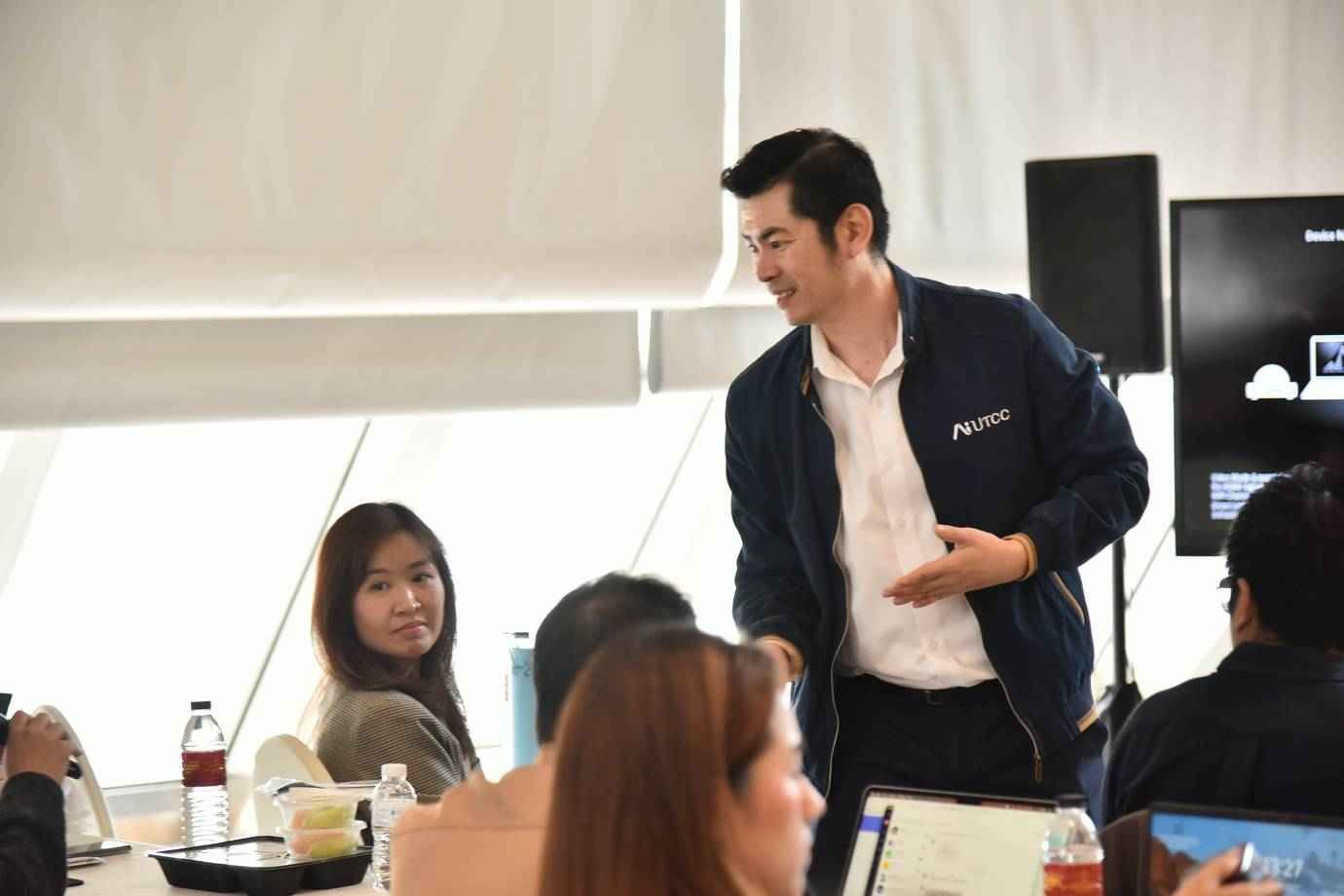 Man in dark jacket gestures, speaking to colleagues at a bright conference table.