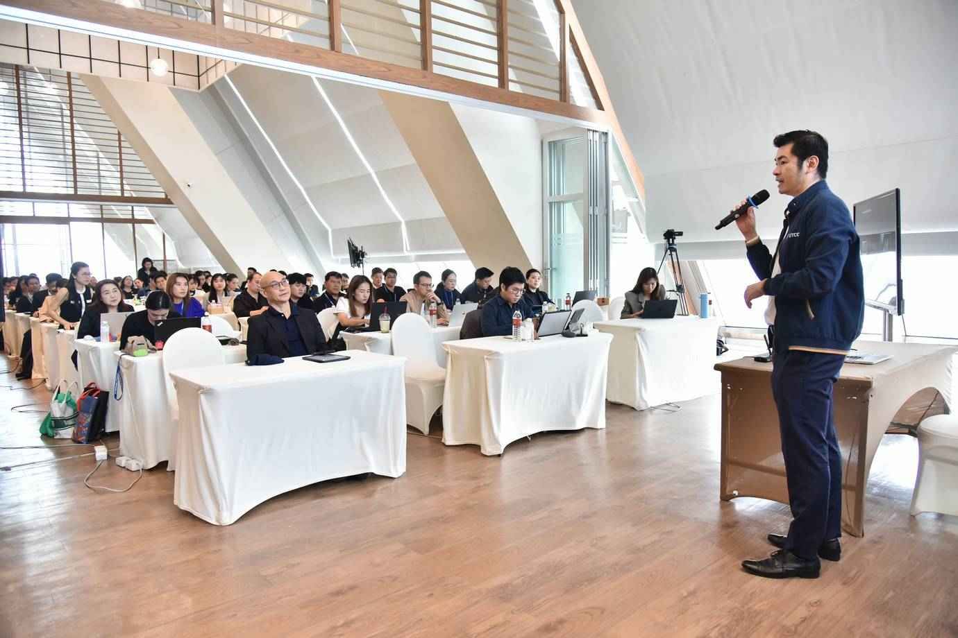 A man in a suit speaks into a microphone before an audience at a conference.