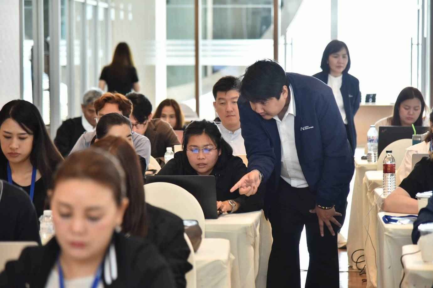 Man assists woman at laptop during a professional training session with other attendees.