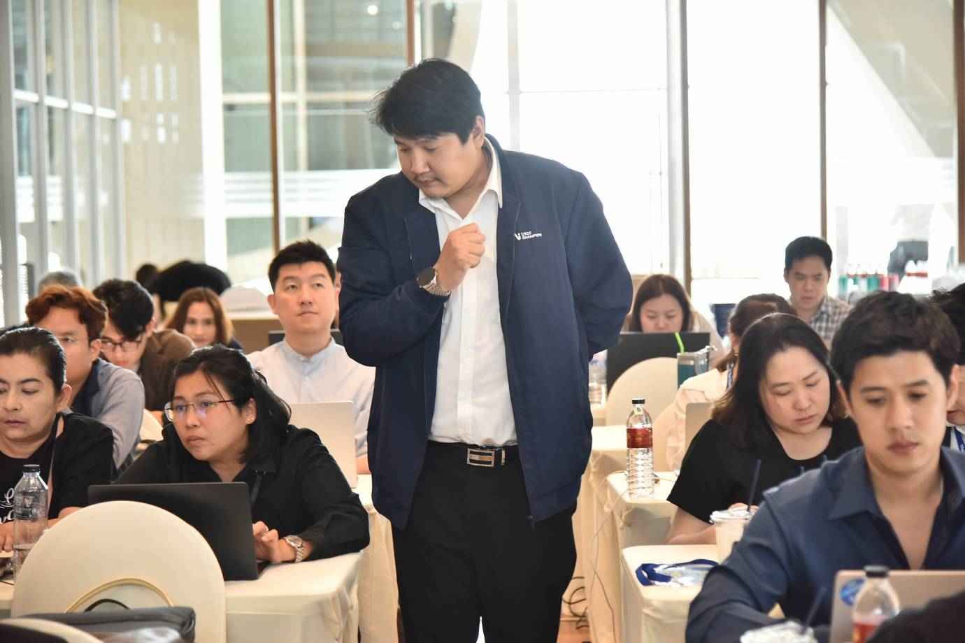 A man in a dark jacket addresses an audience seated at tables during a workshop.