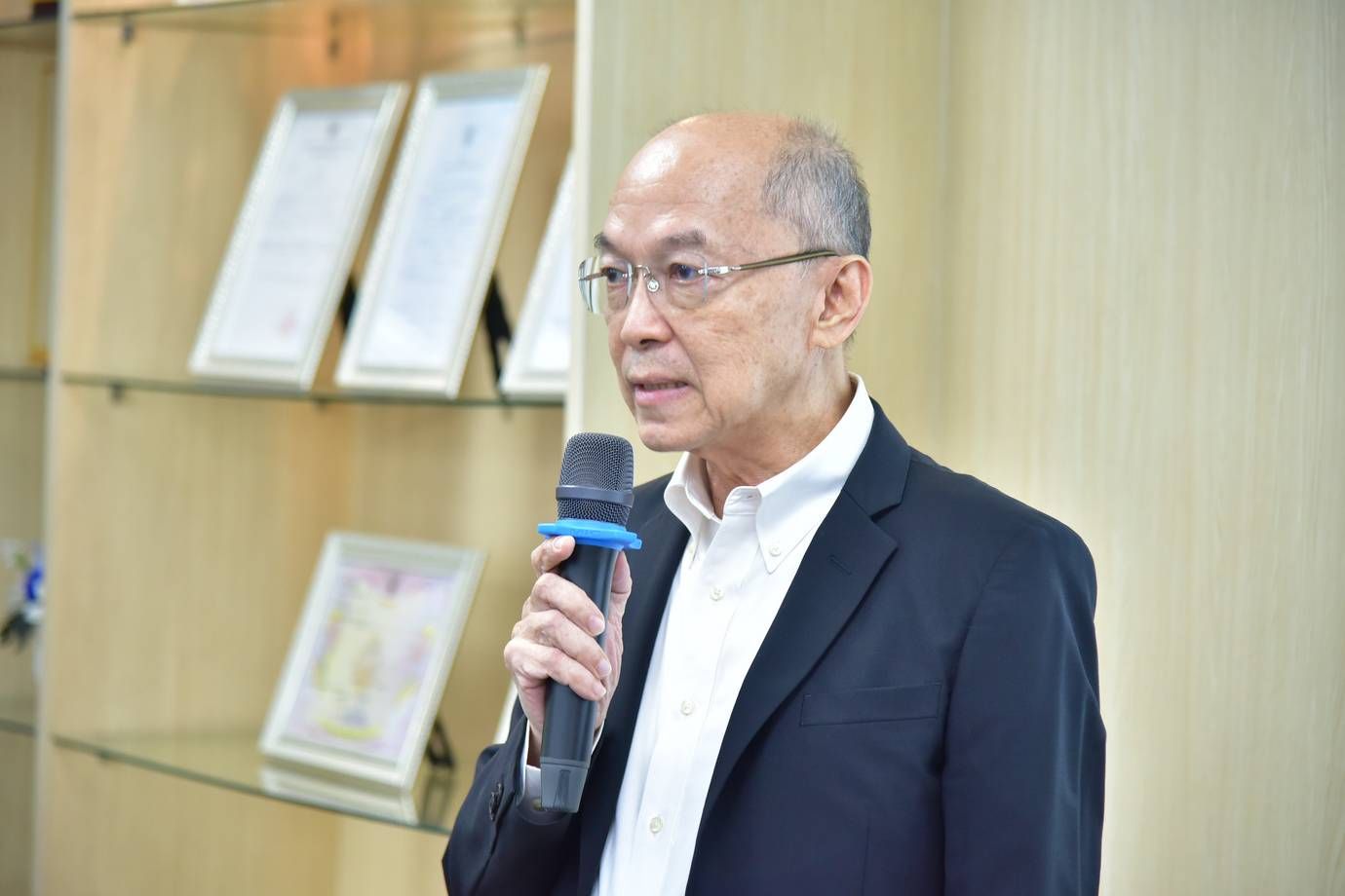 Man in glasses speaks into a microphone during a formal presentation with documents visible.