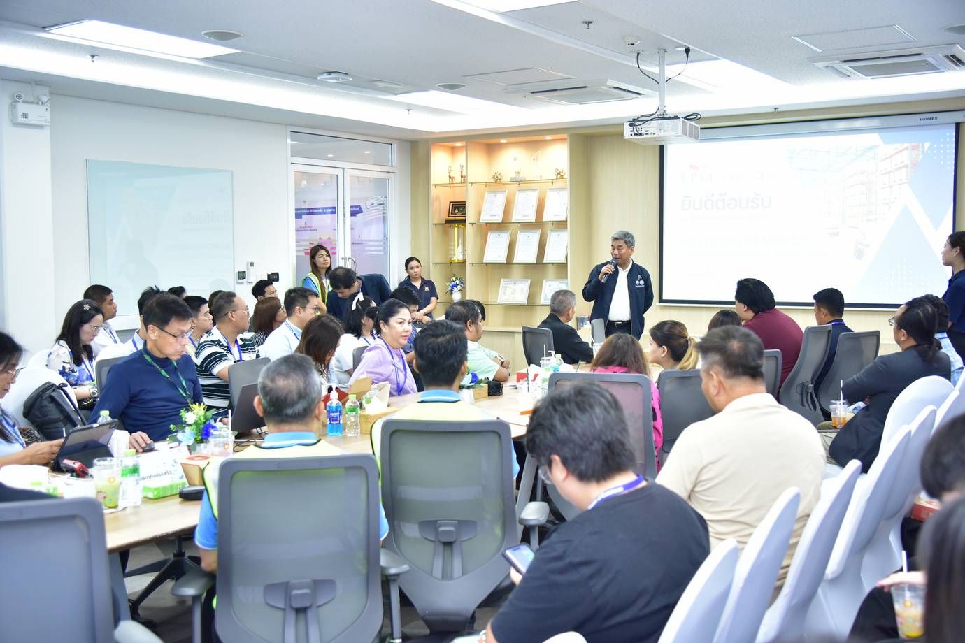 A speaker presents to a seated audience in a bright conference room with a projector.