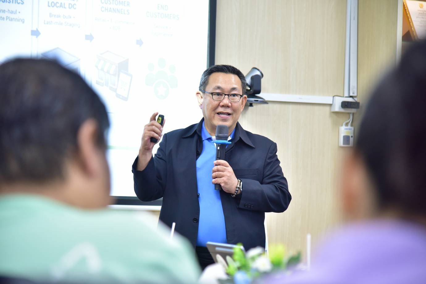 Man in glasses and suit jacket speaking into a microphone during a presentation to an audience.