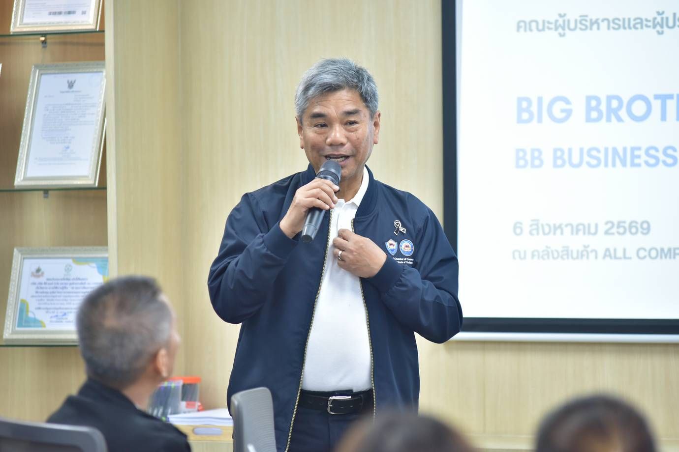 Man speaking into microphone during a business presentation, with 'BIG BROTHER BUSINESS' on screen.
