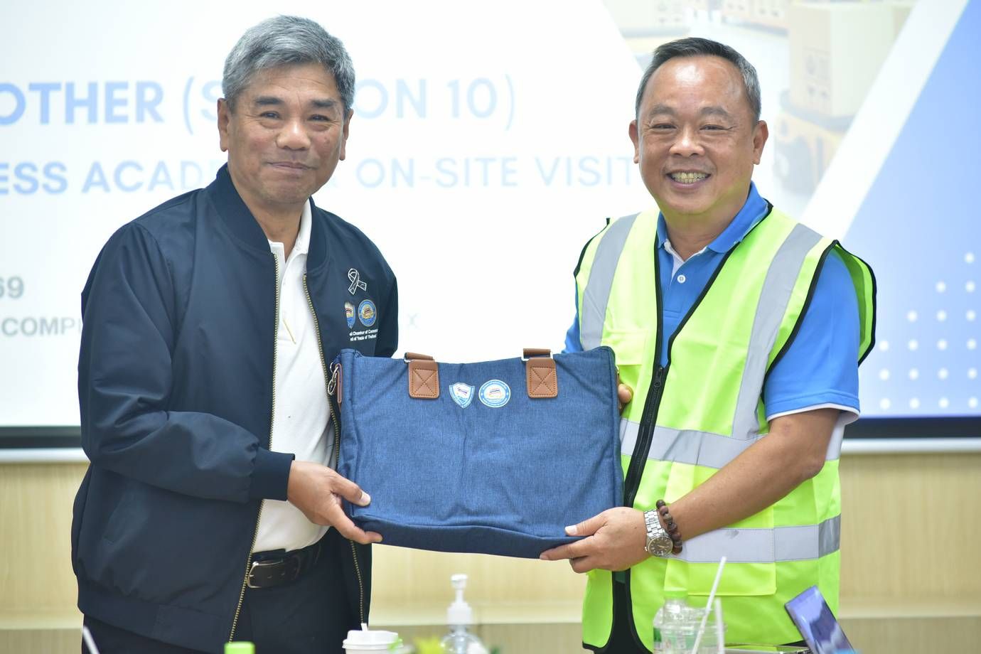 Two smiling men exchange a blue gift bag during a professional event indoors.