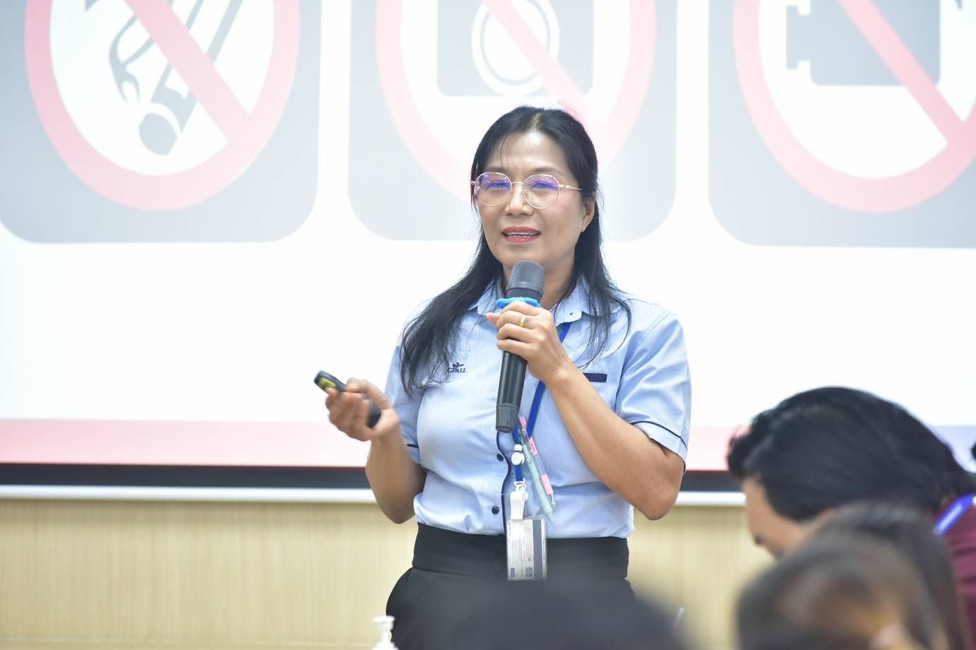 Woman in glasses speaking into a microphone, holding a clicker during a presentation.
