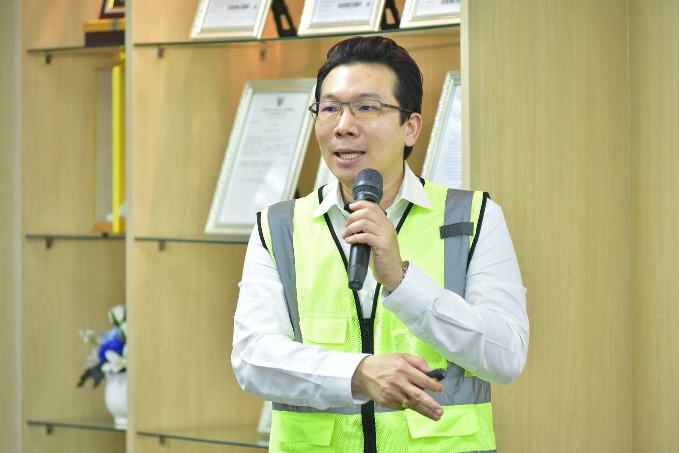 Man in yellow safety vest and glasses speaking into a microphone at an event.