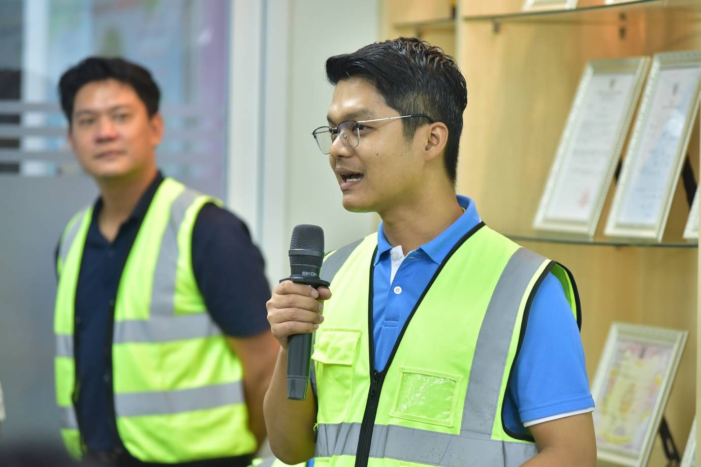 Man in blue shirt and yellow safety vest speaks into microphone indoors.