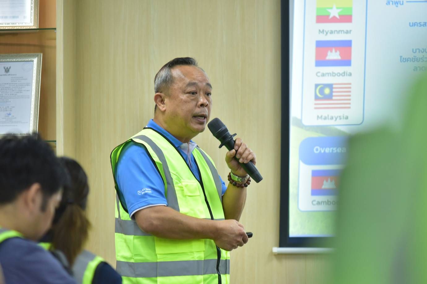 Man in safety vest speaks into microphone during presentation with flags on screen.