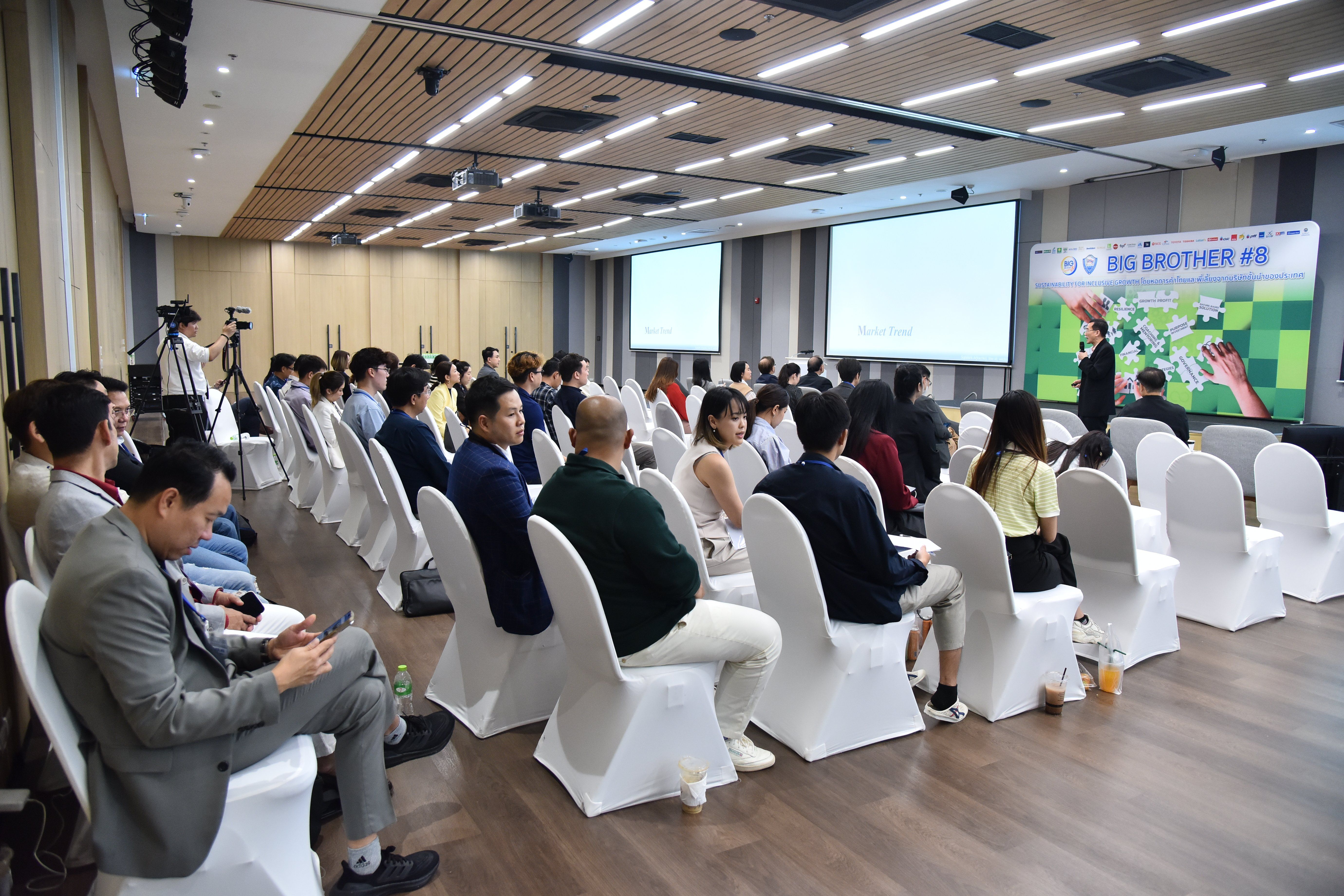 Audience watches a presenter at a seminar, seated in a well-lit conference room.