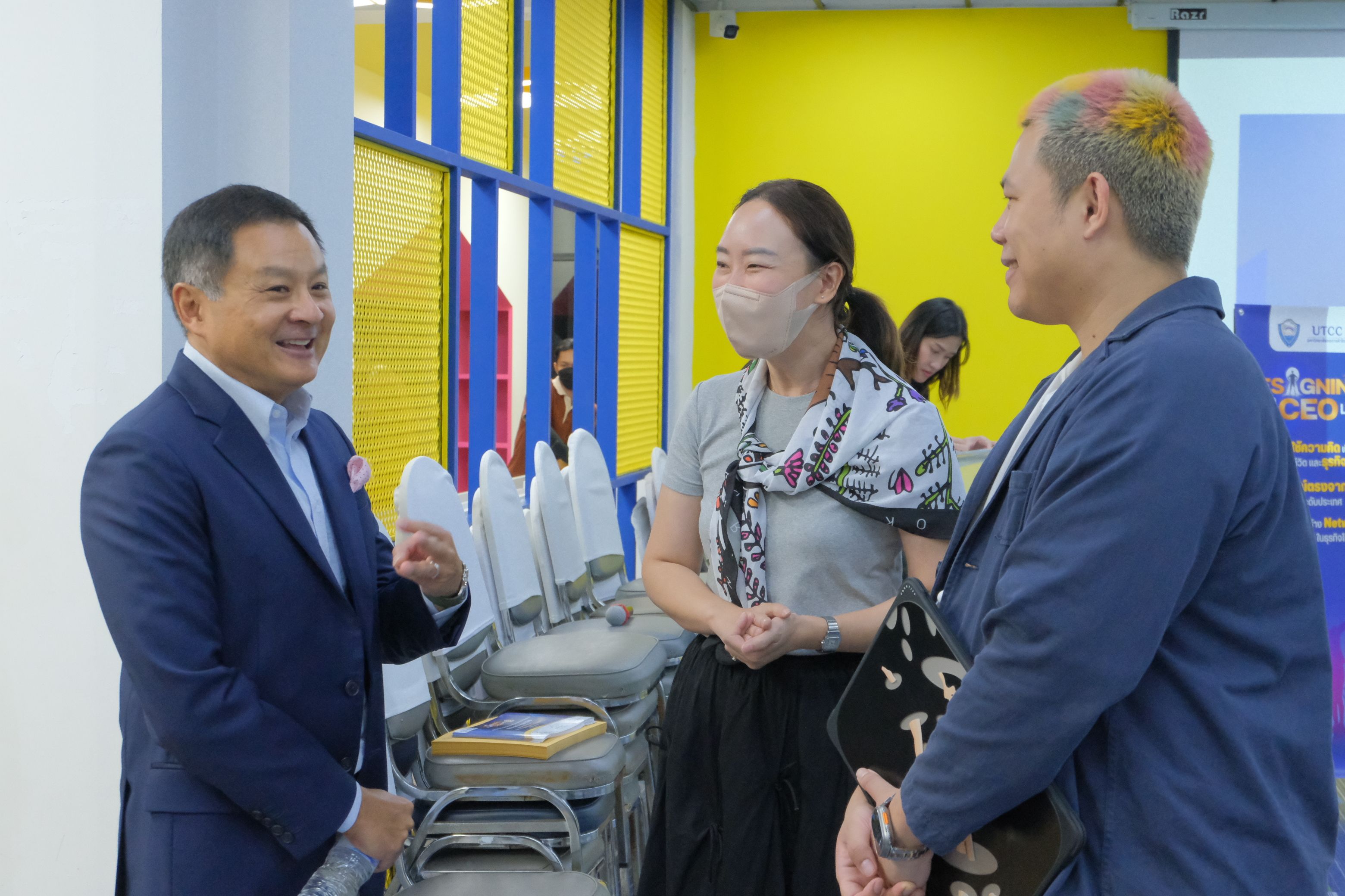 Three professionals, two men and one masked woman, converse in a modern yellow-walled room.