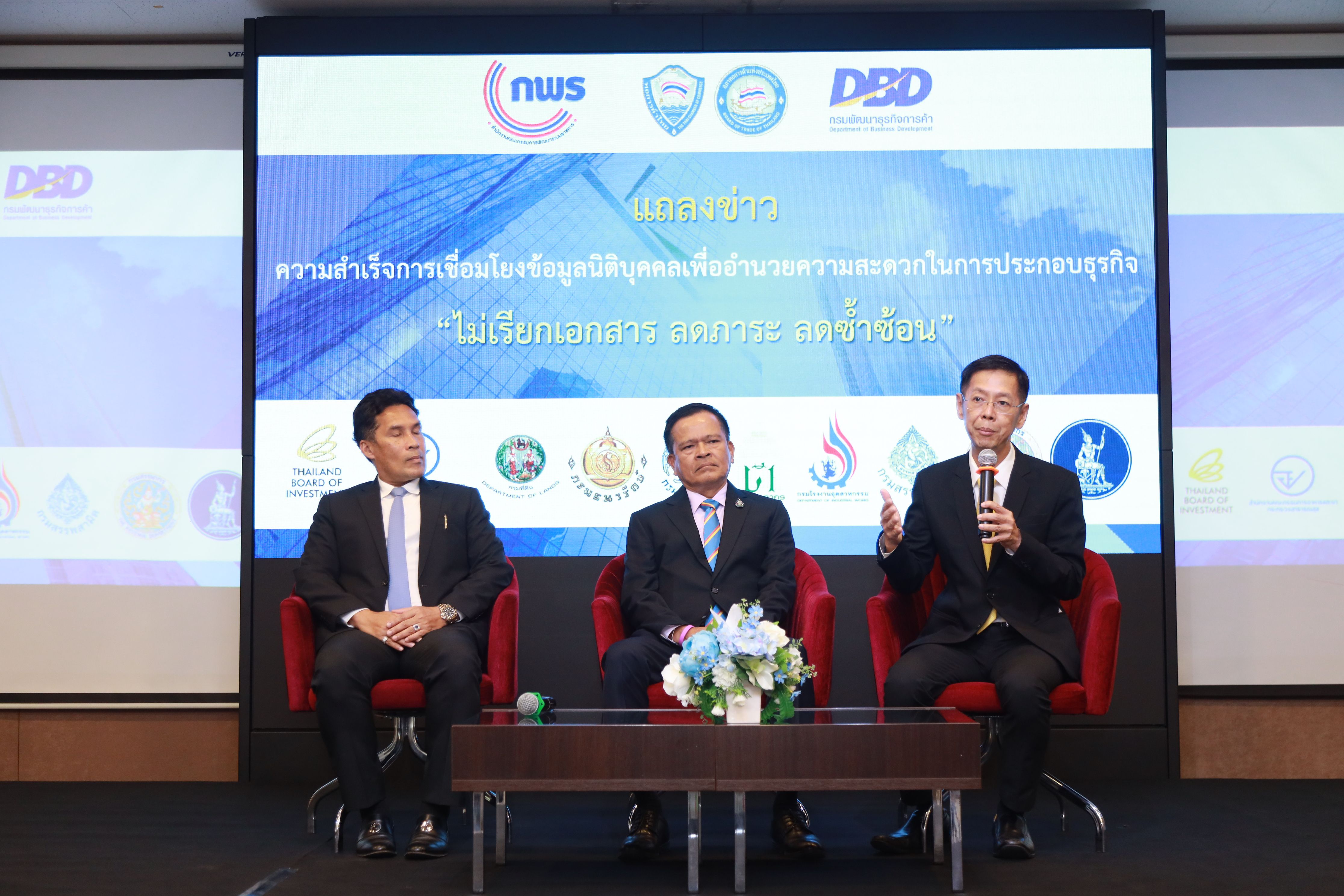 Three men in suits sit on a stage, one actively speaks before a large screen.