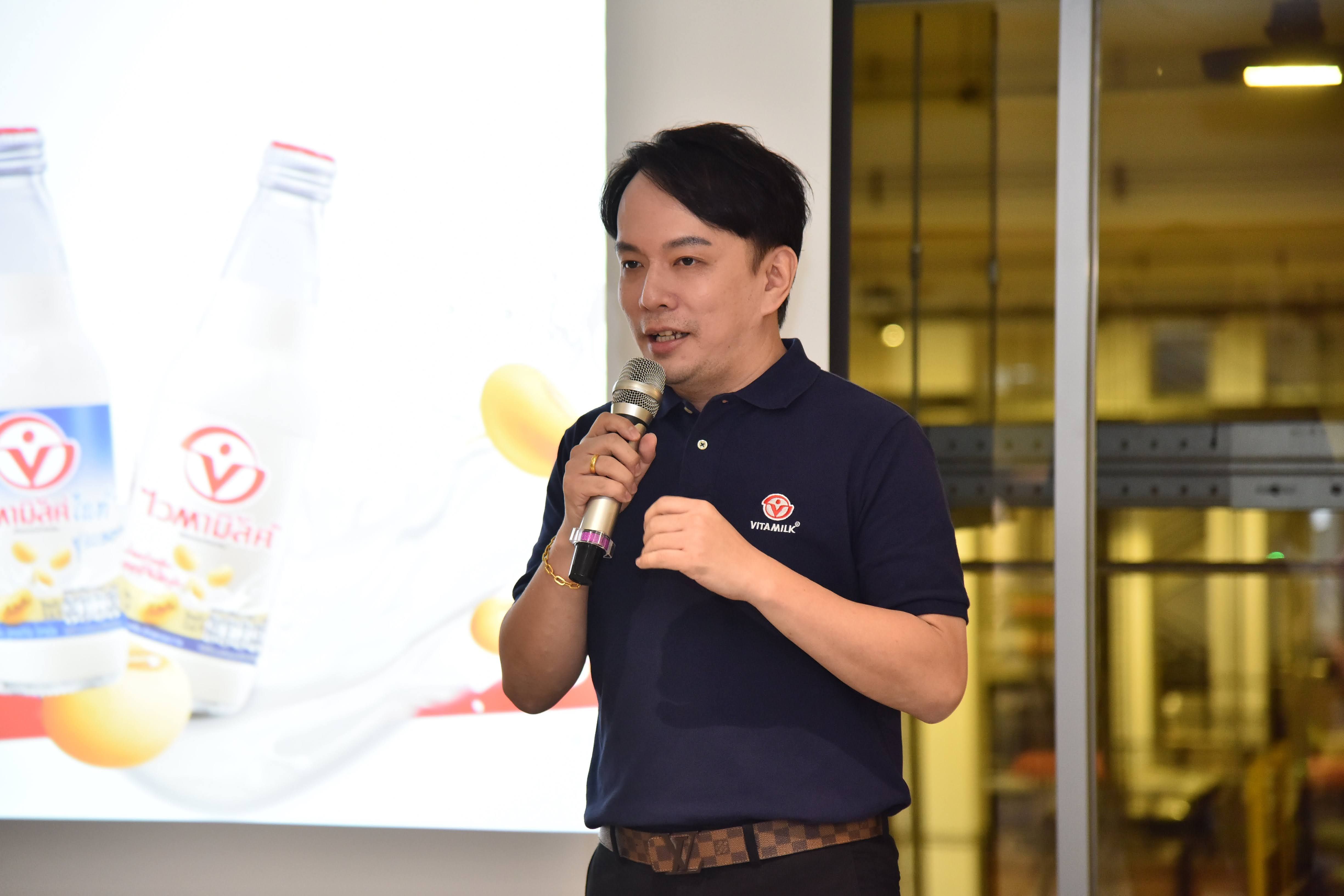 Man speaking into microphone during a presentation featuring bottled beverages and soybeans.