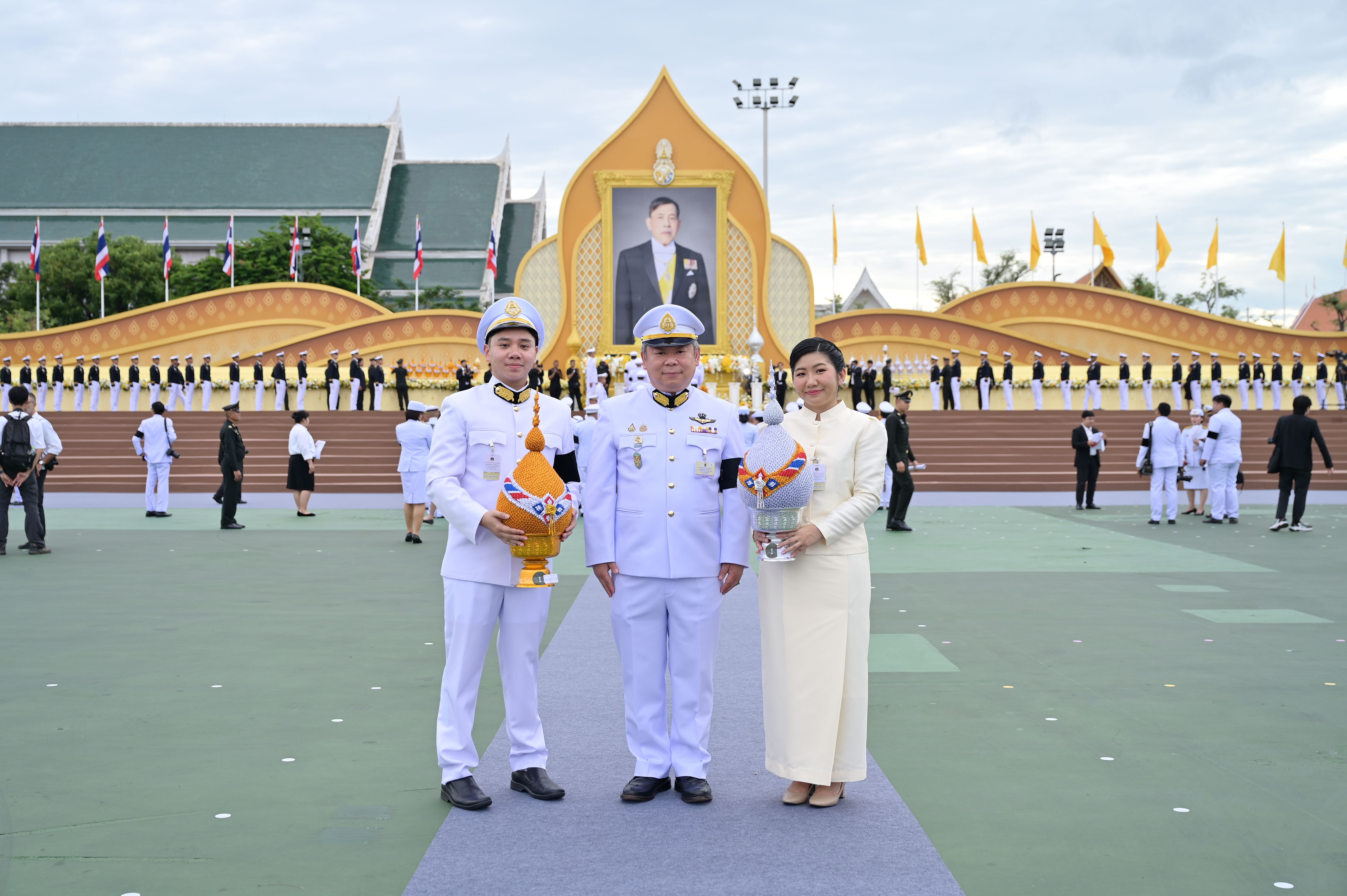 Three people in formal Thai attire stand before a large golden royal portrait.